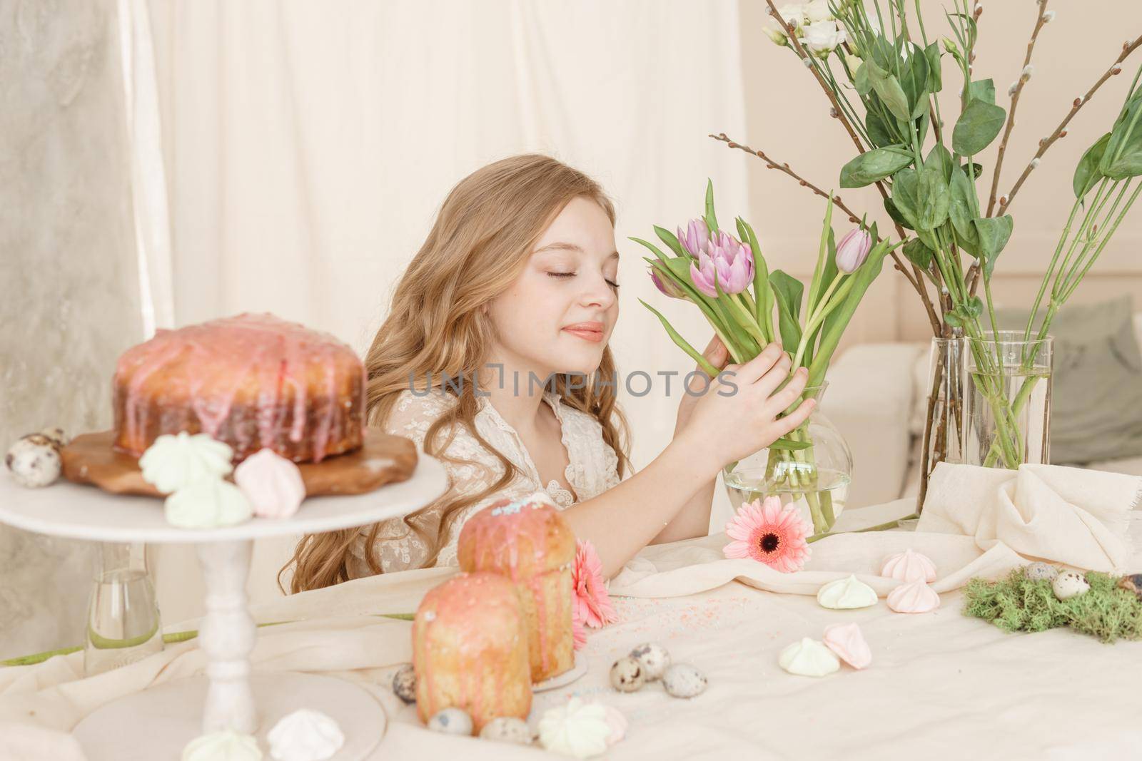 A girl with long hair in a light dress is sitting at the Easter table with cakes, spring flowers and quail eggs. Happy Easter celebration