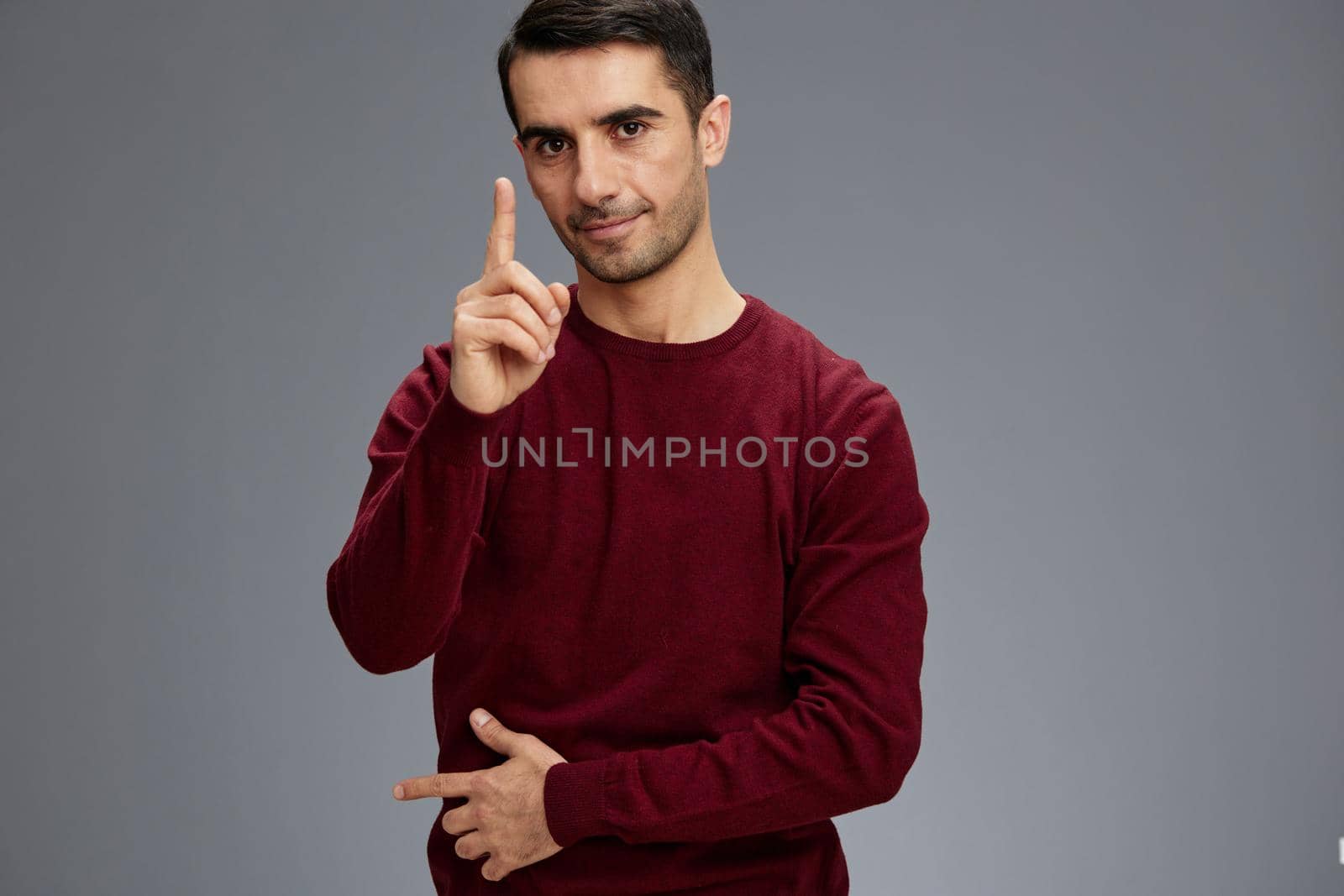 successful man holding index finger near face posing gesture with hands red sweater Gray background by SHOTPRIME