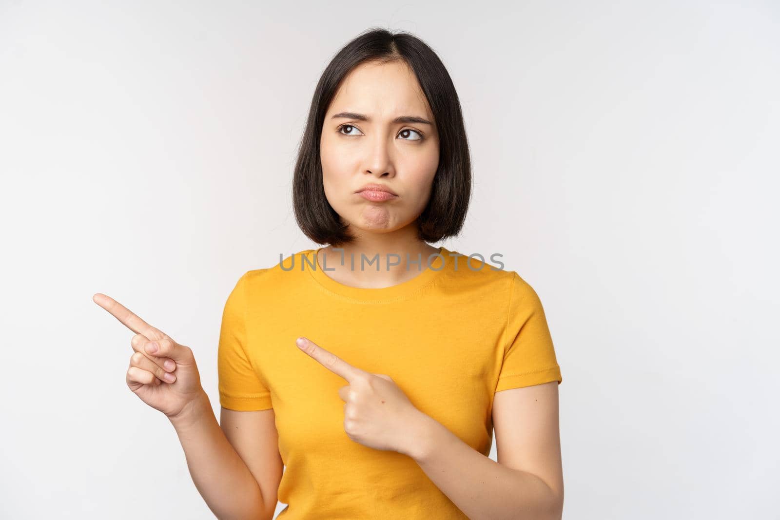 Upset grumpy asian girl, pouting and looking at smth unfair, pointing fingers left at banner, logo brand, standing in yellow tshirt over white background by Benzoix