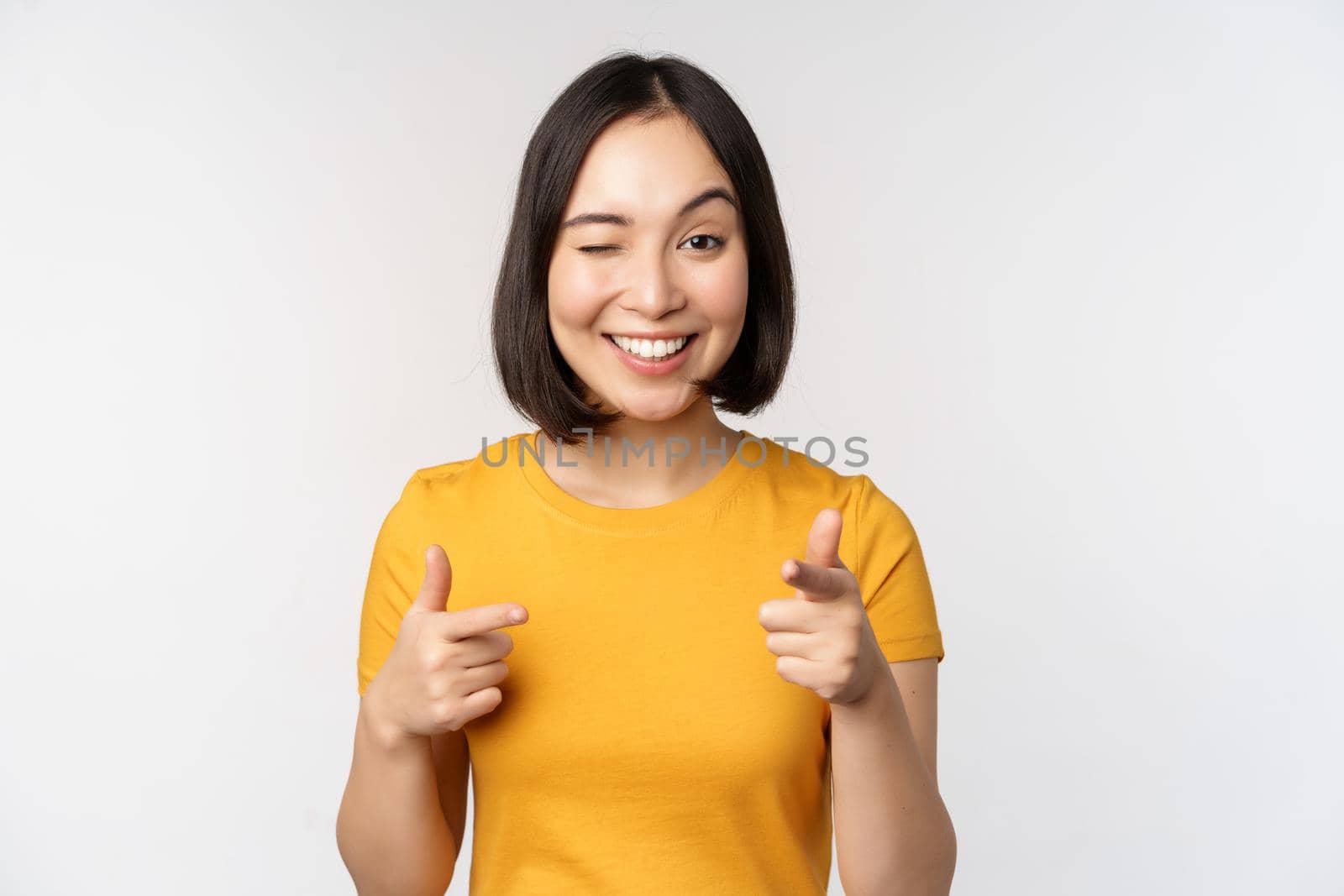 Young korean girl student, pointing fingers at camera and smiling, congratulating you, choosing, inviting people, standing over white background.