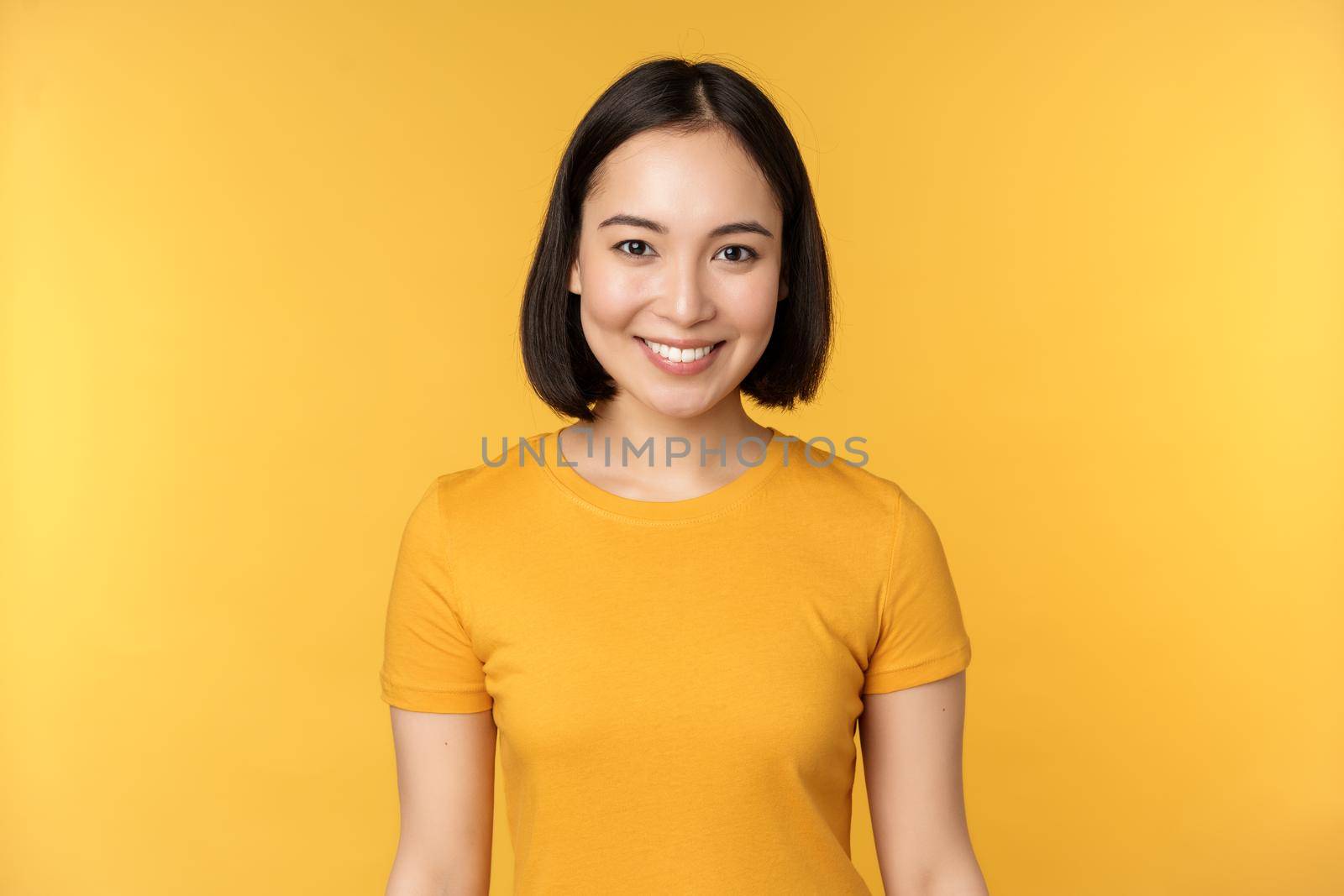 Portrait of young modern asian woman, smiling happy with white teeth, looking confident at camera, wearing casual t-shirt, standing over yellow background.