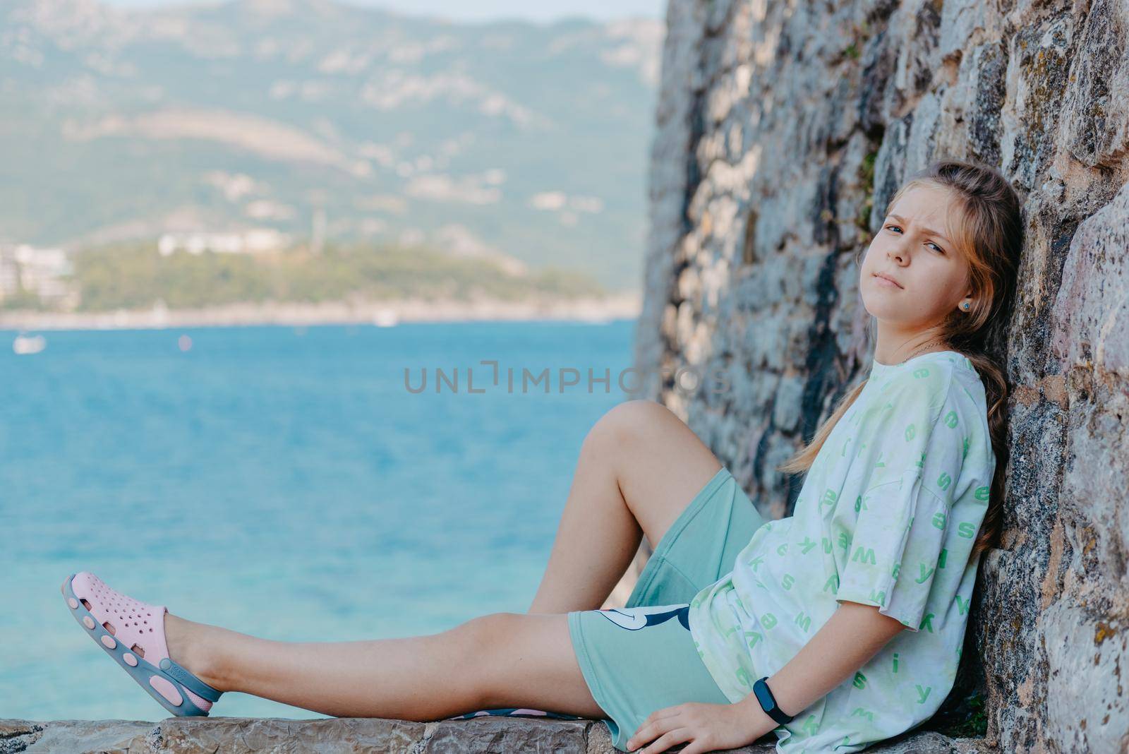 Beautiful girl sitting on a stone wall, in background is the blue sea, Budva, Montenegro. by Andrii_Ko