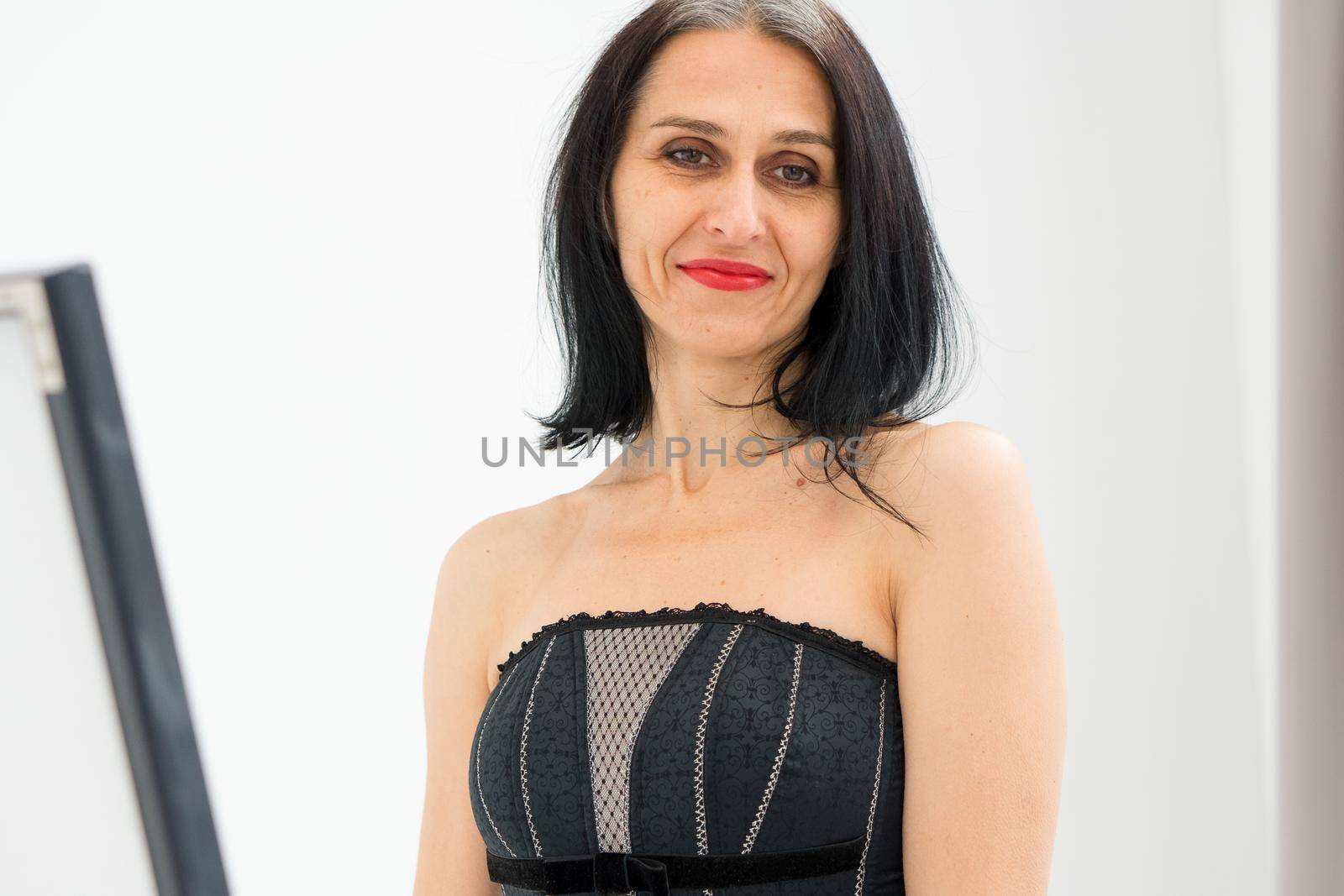 Middle aged woman starting getting grey-haired is posing in studio in black clothes and stockings on white background, middle age sexy lady, cosmetology concept.
