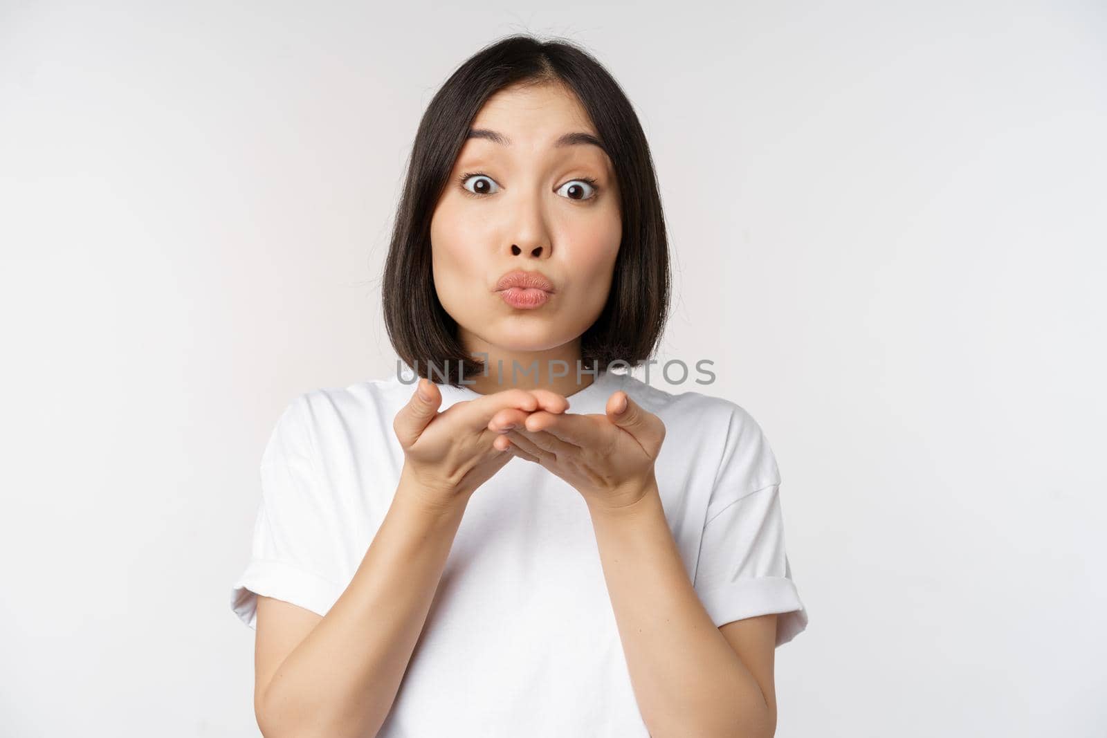 Lovely young asian woman, sending air kiss and looking coquettish at camera, standing in tshirt over white background by Benzoix