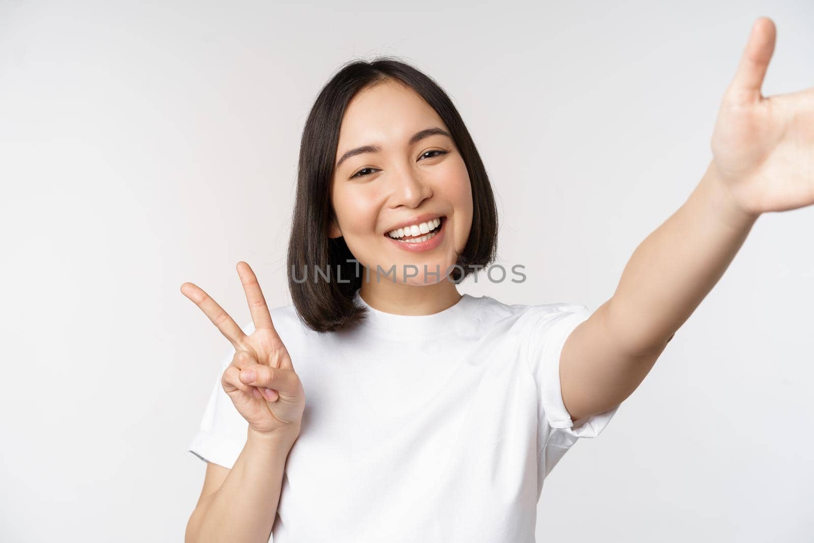 Beautiful young asian woman taking selfie, posing with peace v-sign, smiling happy, take photo, posing against white background by Benzoix