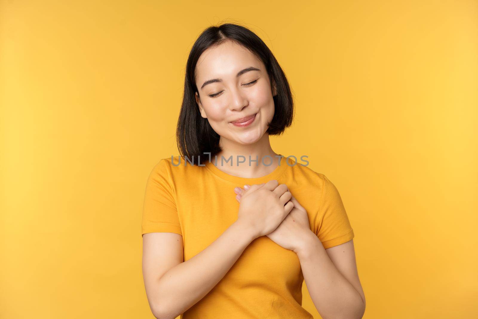 Romantic korean girl, asian woman holding hands on heart, smiling with care and tenderness, standing over yellow background by Benzoix