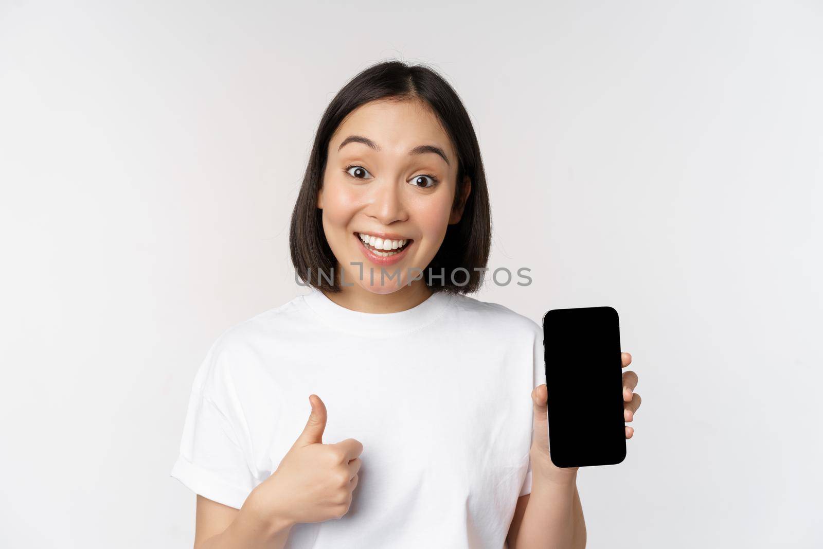 Enthusiastic young woman showing thumb up and mobile phone screen, standing in tshirt over white background by Benzoix