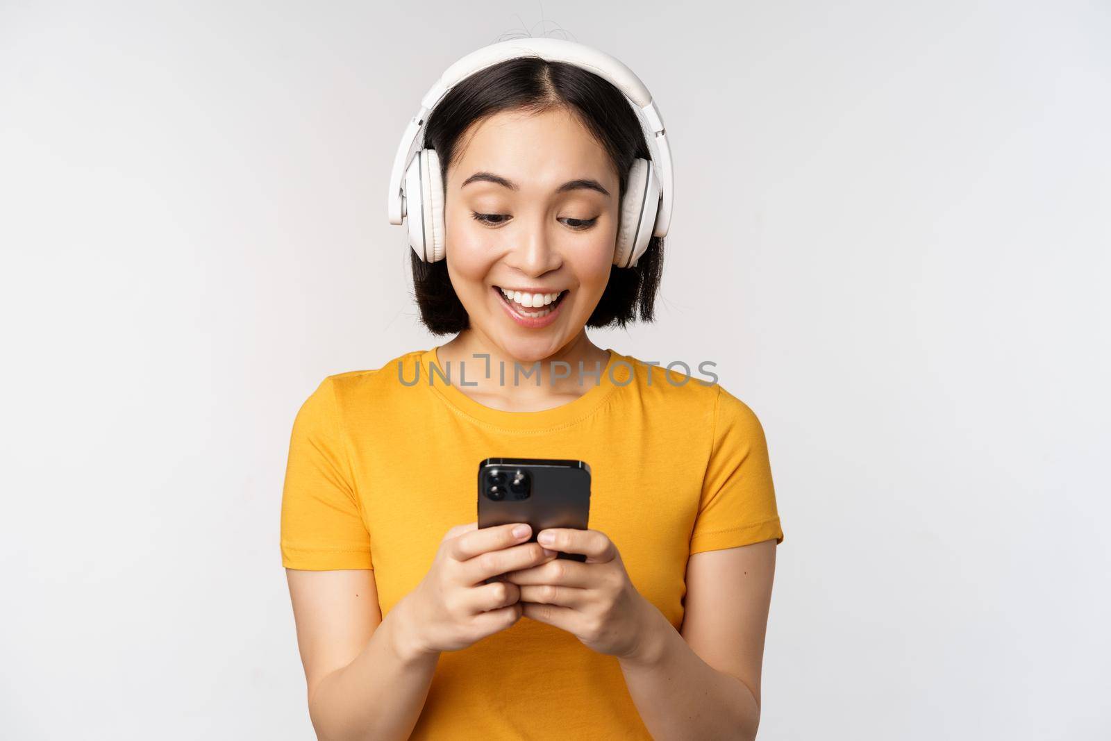 Cute japanese girl in headphones, looking at mobile phone and smiling, using music app on smartphone, standing against white background.