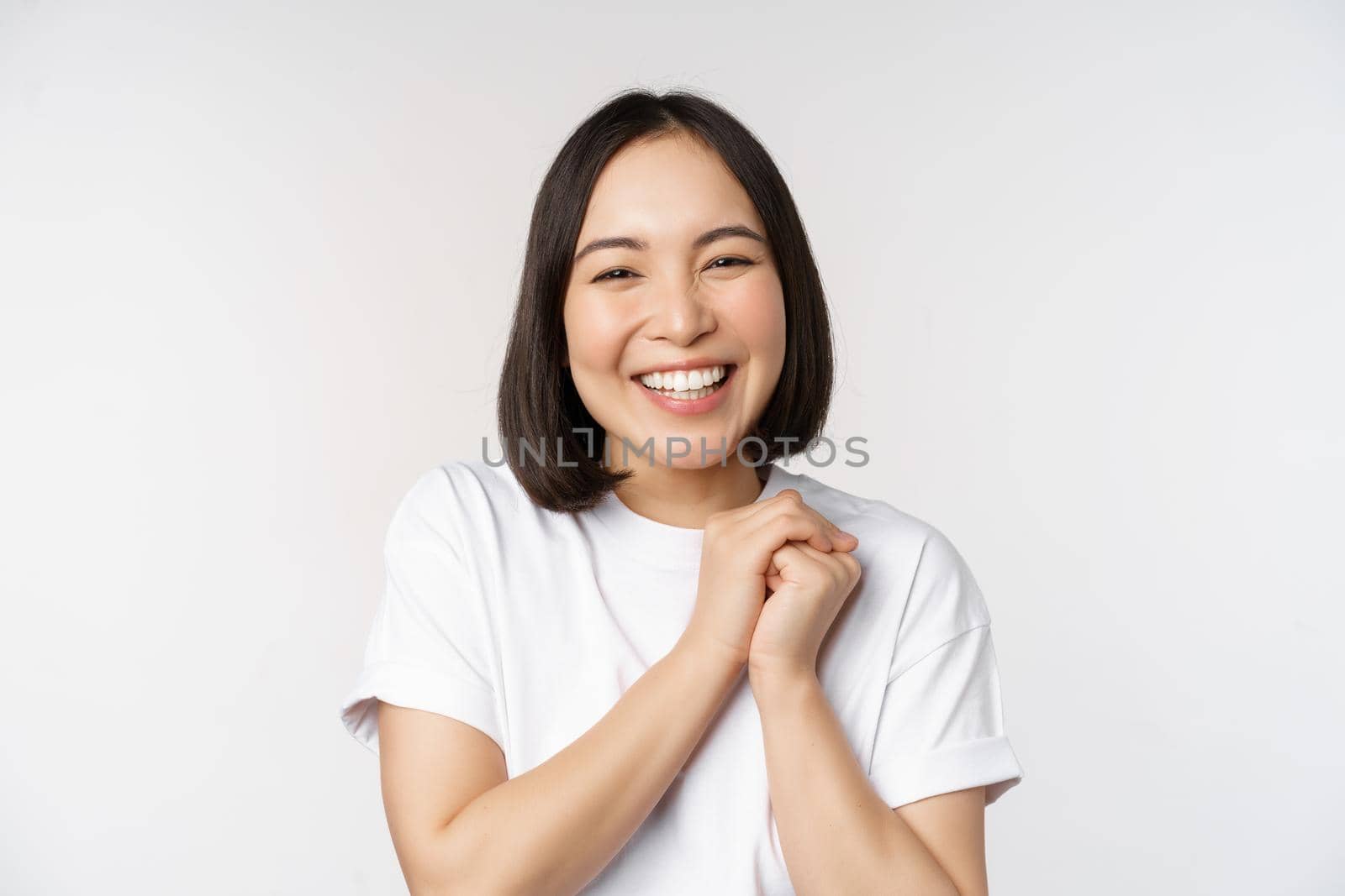 Portrait of beautiful korean woman with healthy white smile, laughing and looking happy at camera, standing in tshirt over white studio background.
