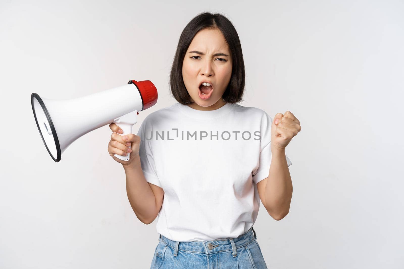 Angry asian girl activist, holding megaphone and looking furious, protesting, standing over white background. Copy space