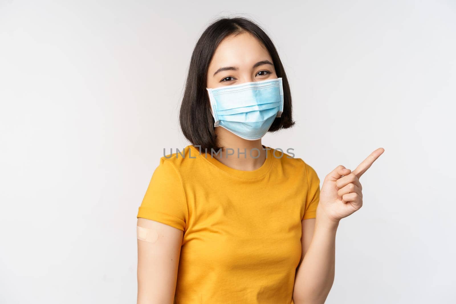 Covid-19, vaccination and healthcare concept. Portrait of cute asian girl in medical mask, has band aid on shoulder after coronavirus vaccine, standing over white background.