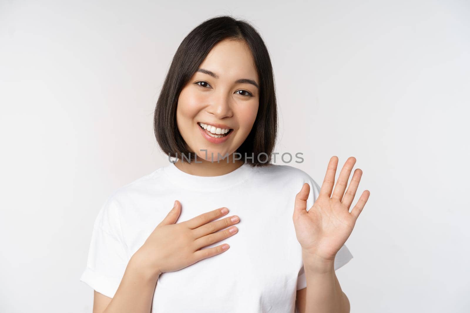 Portrait of beautiful korean girl raising hand, introduce herself, put hand on heart, greeting, standing over white background.