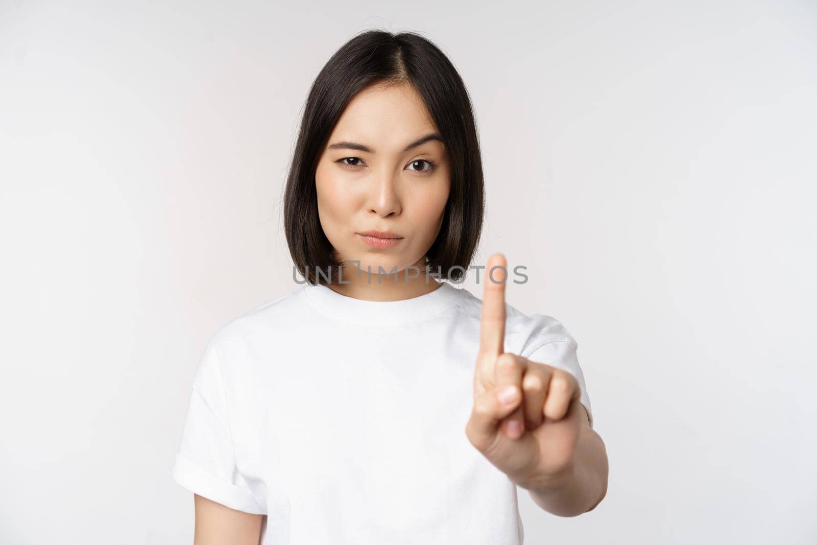Image of asian girl showing stop, prohibit smth, extend one arm to show forbidding, taboo gesture, standing in tshirt over white background.