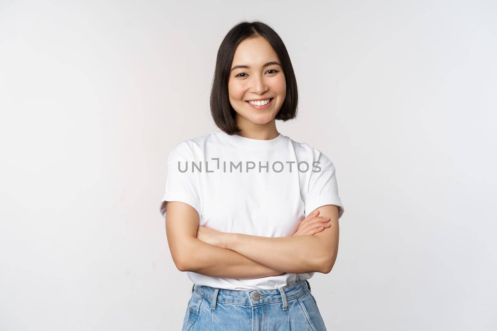 Portrait of happy asian woman smiling, posing confident, cross arms on chest, standing against studio background.