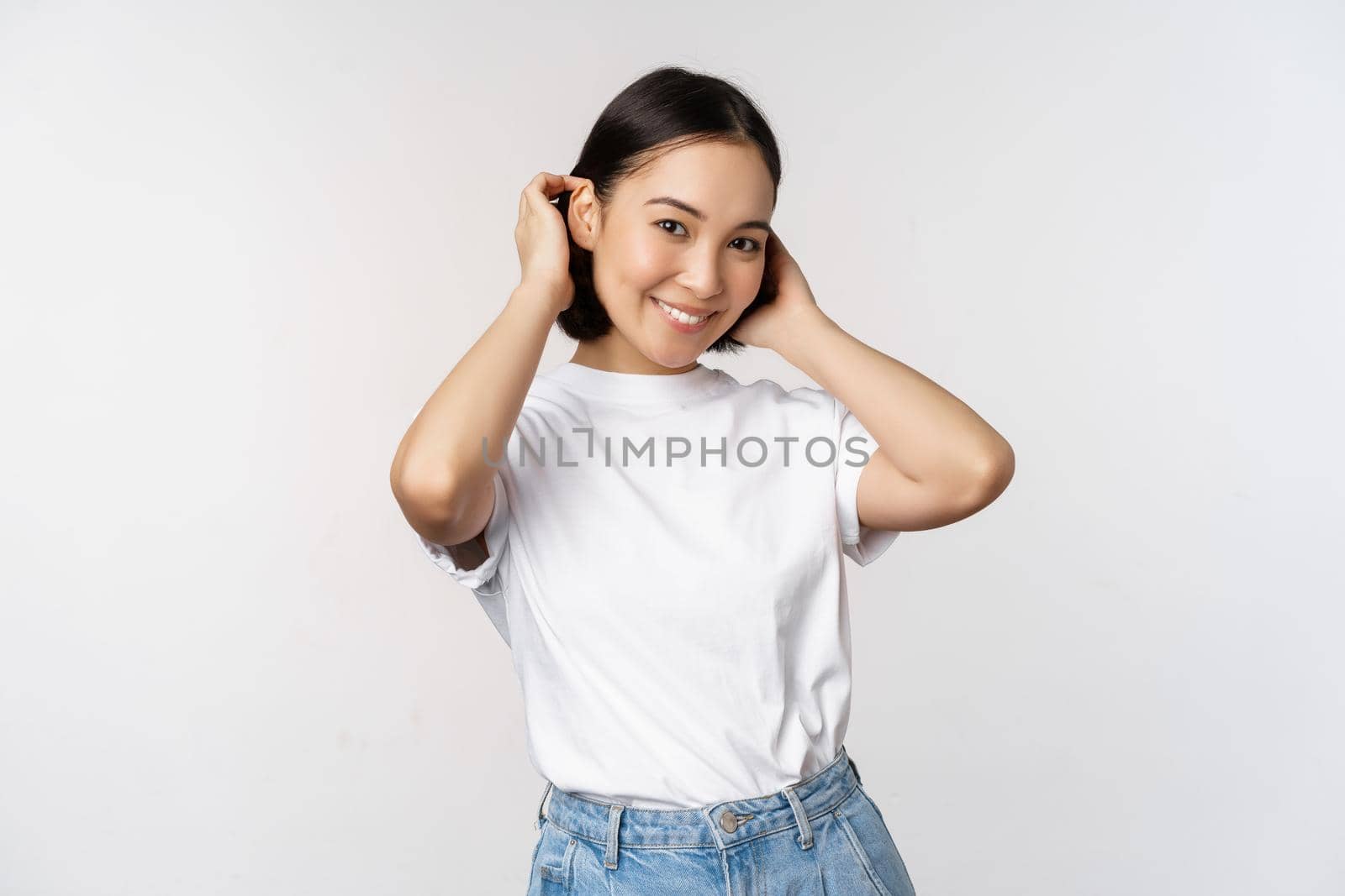 Portrait of cute, beautiful asian woman touching her new short haircut, showing hairstyle, smiling happy at camera, standing over white background.