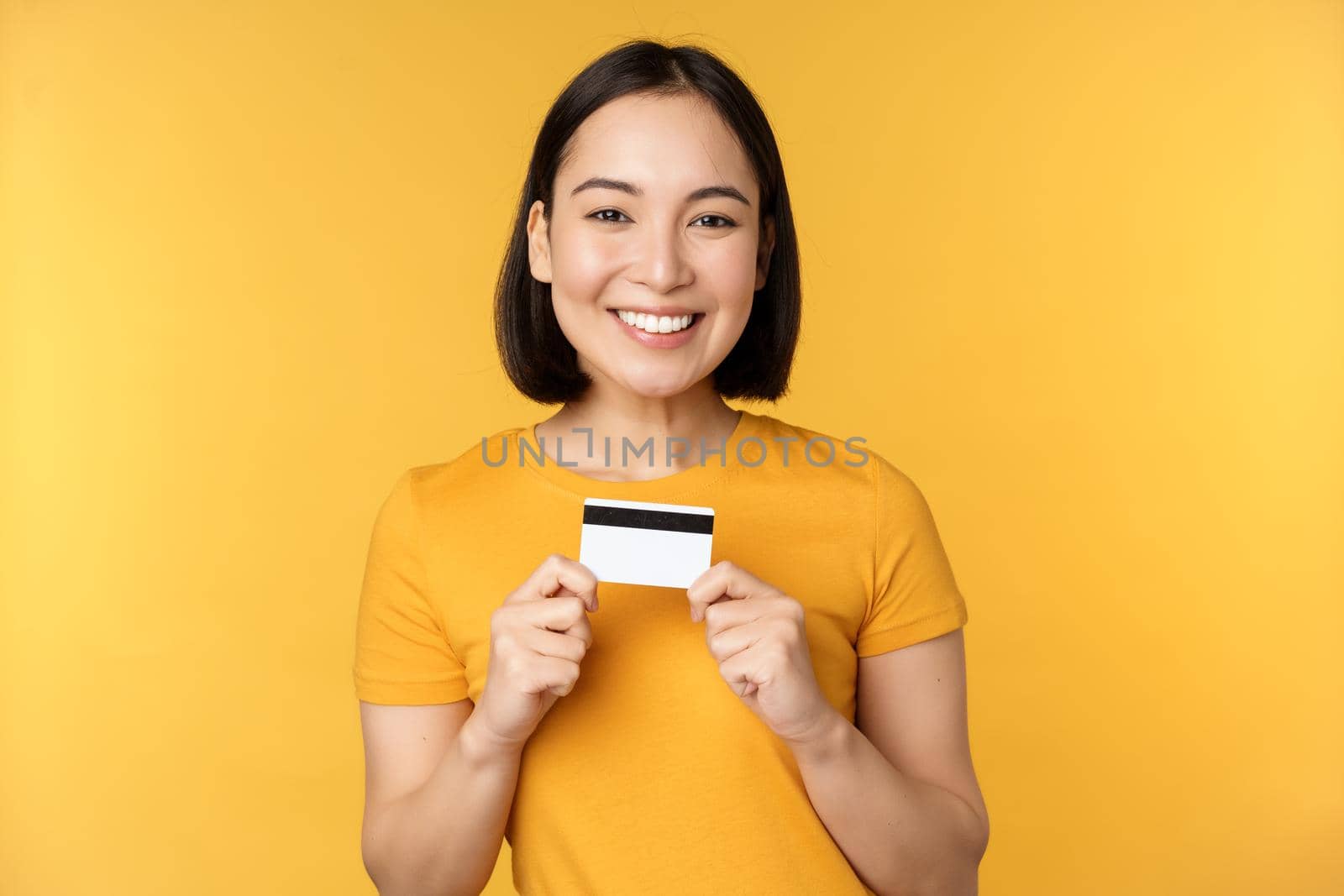 Beautiful asian woman showing credit card and smiling, recommending bank service, standing over yellow background by Benzoix