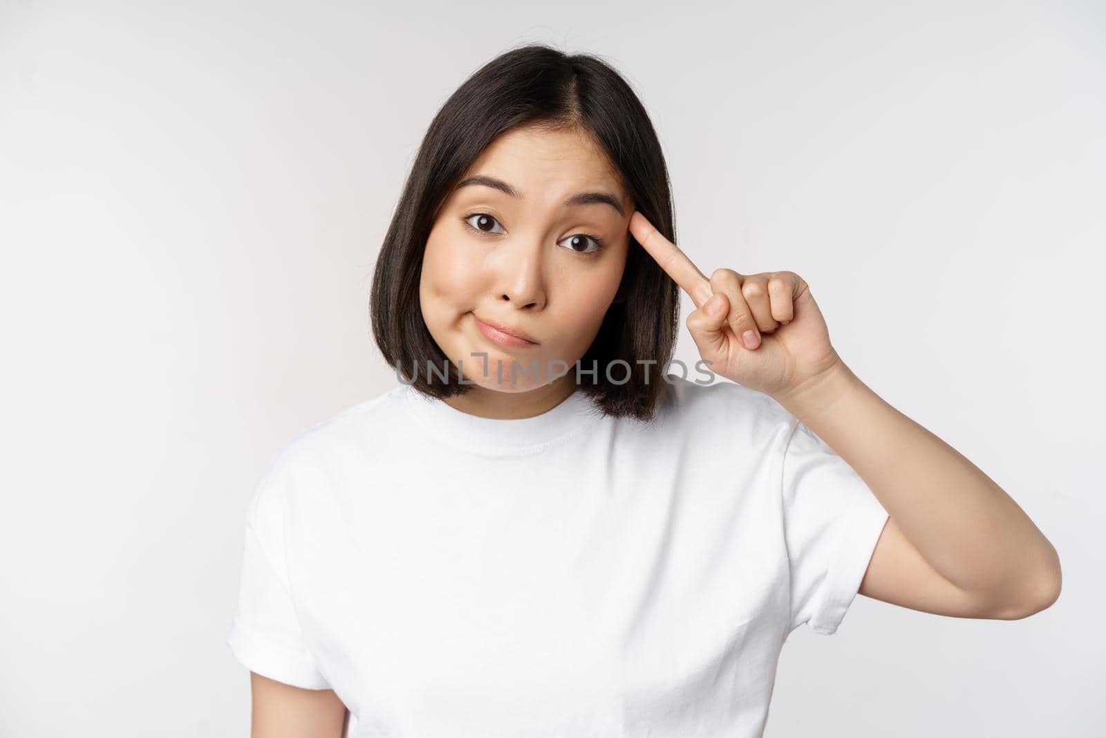 Are you crazy. Young asian woman roll finger on head, mocking someone stupid, standing in tshirt over white background. Copy space