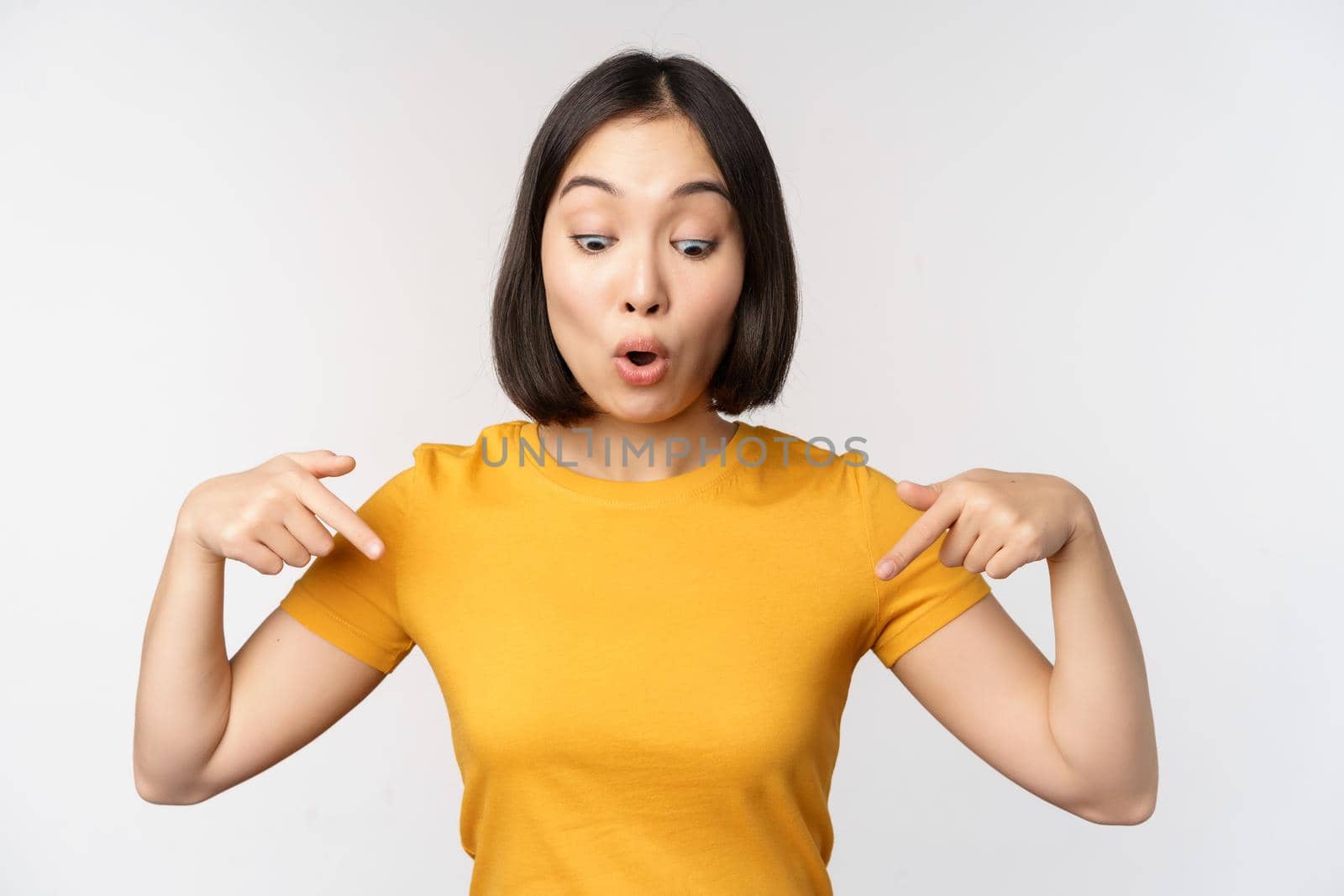 Happy asian girl pointing fingers down, looking at announcement, banner or advertisment, standing in yellow tshirt over white background.