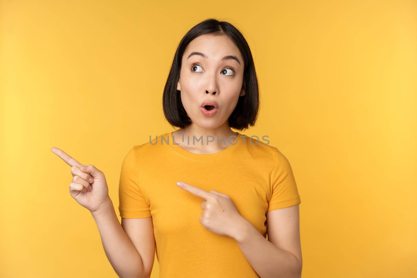 Portrait of intrigued asian woman, looking and pointing fingers left at advertisement, showing smth interesting, standing over yellow background.