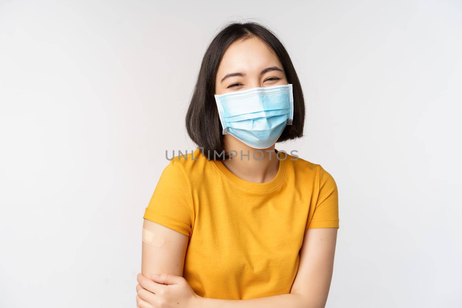 Healthy and happy asian woman in medical face mask, has band aid on shoulder after covid-19 vaccination, got vaccinated from omicron delta variant, whtie background by Benzoix