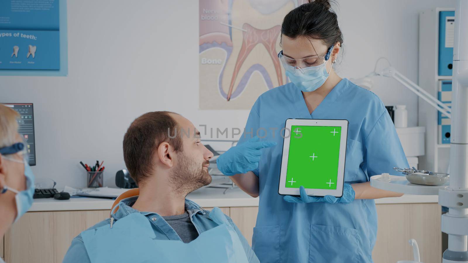 Assistant vertically holding digital tablet with green screen at appointment with patient in cabinet. Woman pointing at mock up template with blank copy space and isolated chroma key.