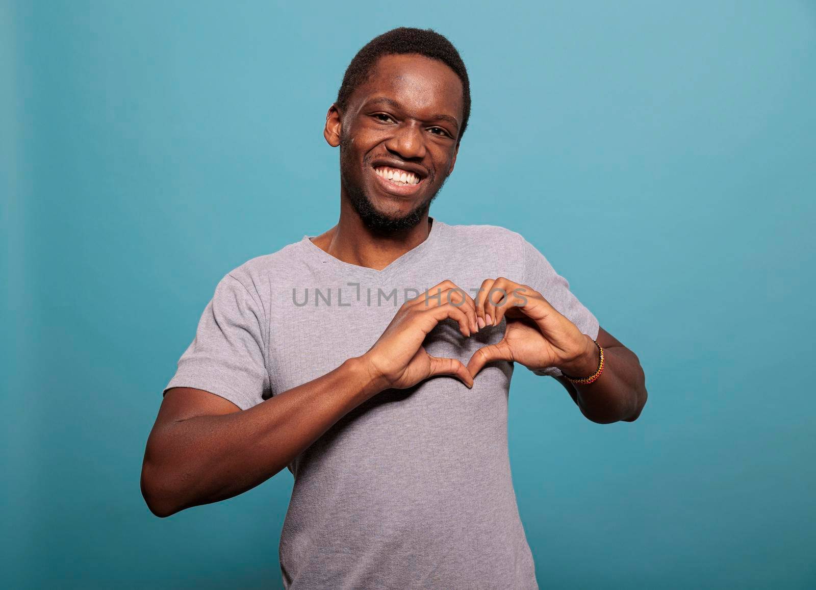 Young adult doing heart shaped sign with hands in studio, expressing amorous feelings on camera. Smiling man doing romantic gesture to express love, affection and passion for valentines day.