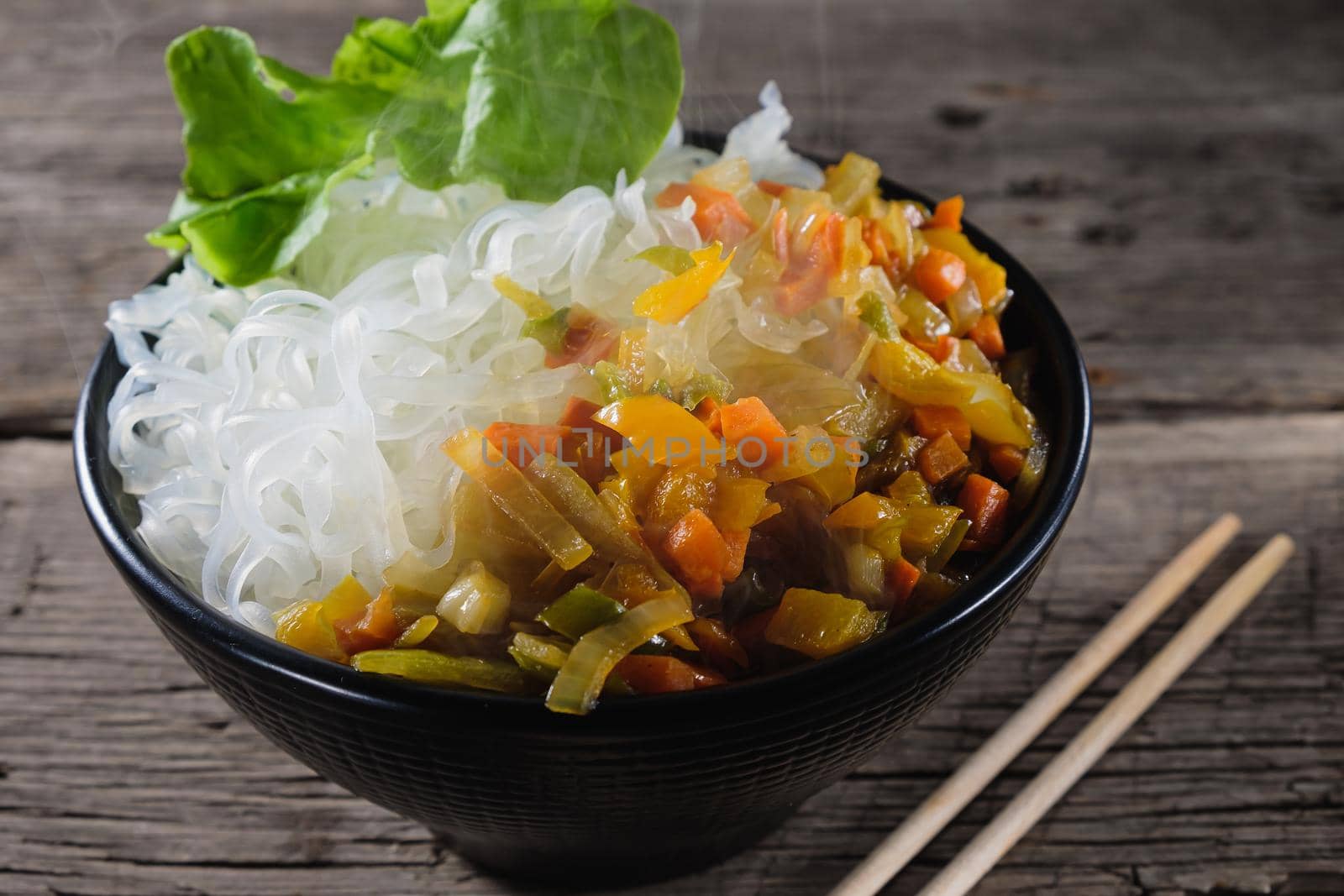 A plate of rice noodles stands on an old wooden table top. Rice noodles are most common in the cuisines of East, Southeast Asia and South Asia. The side dish consists of fried onions, carrots, paprika. Copy space. Horizontal orientation