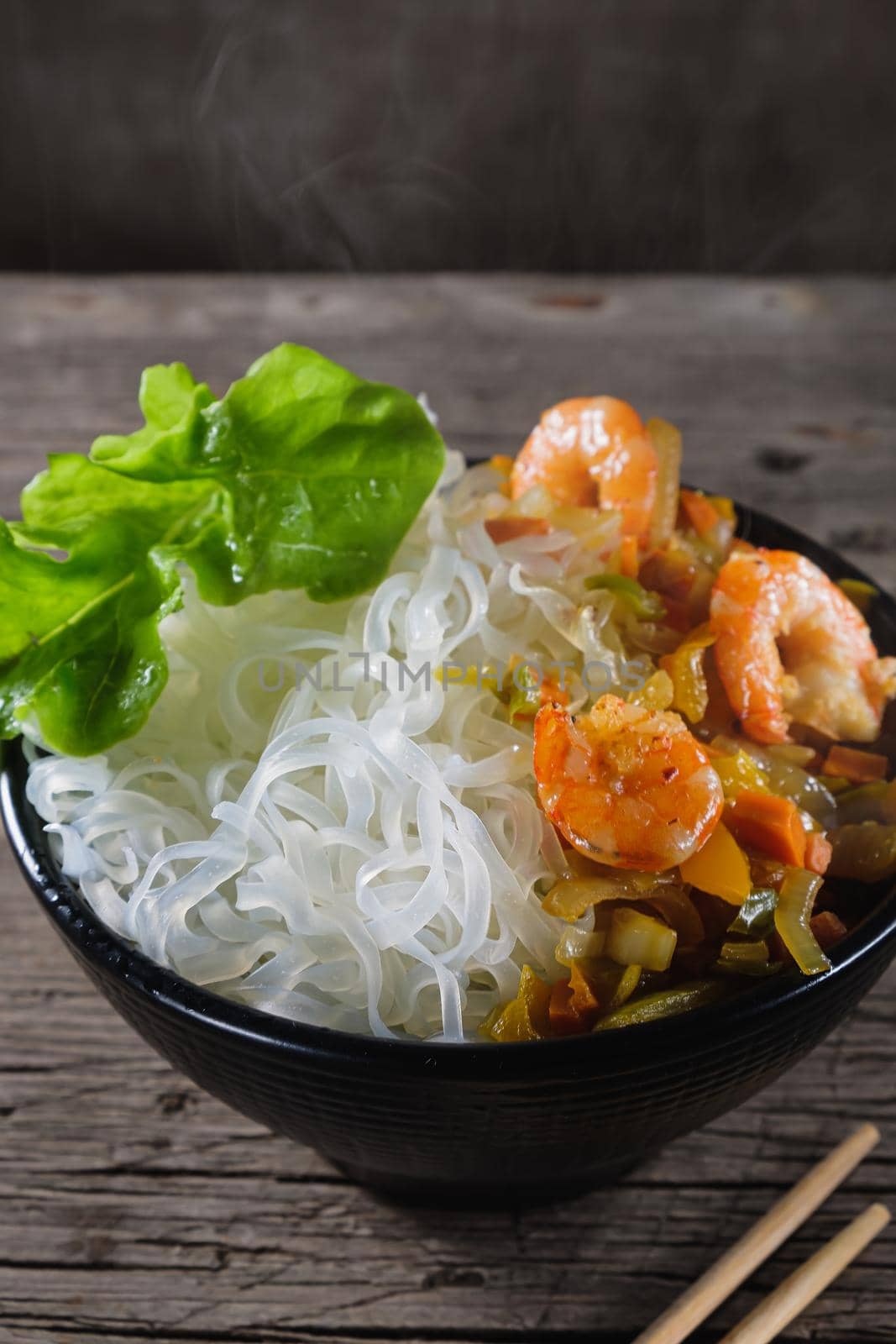 A plate of rice noodles stands on an old wooden table top. Rice noodles are most common in the cuisines of East, Southeast Asia and South Asia. The side dish consists of fried onions, carrots, paprika, shrimp. Copy space. Vertical orientation