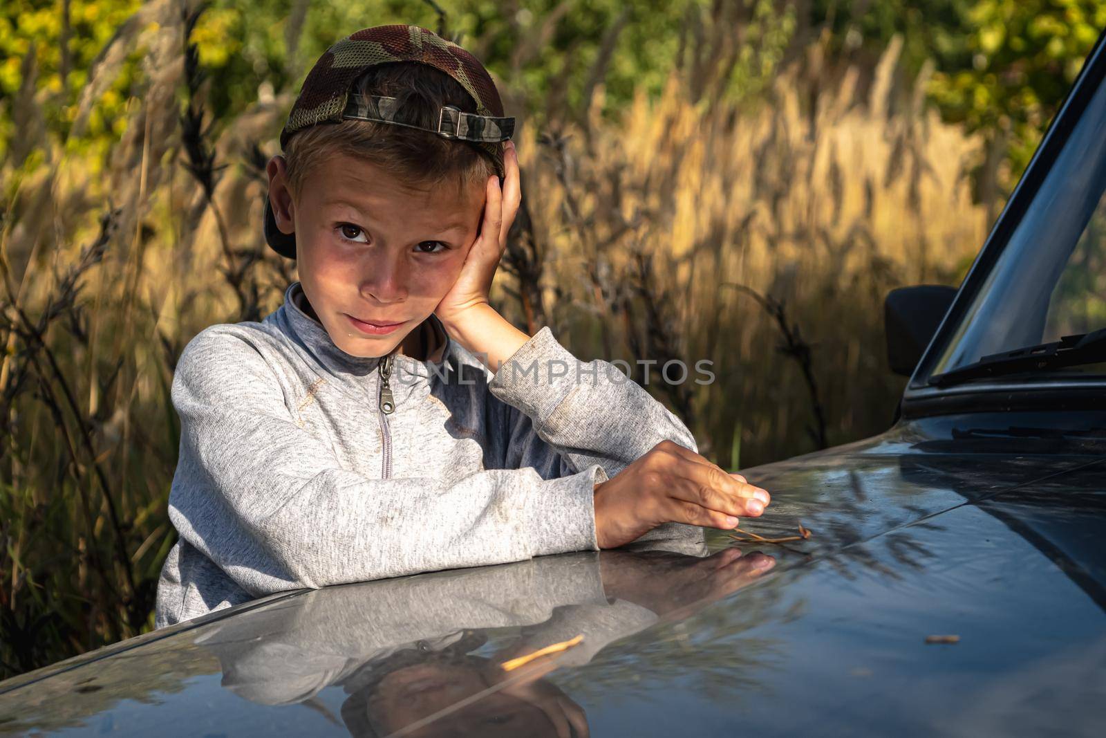 a cute six year old boy stands by the car against the background of autumn grass.. horizontal orientation.