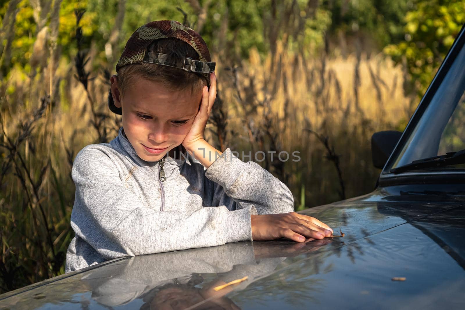 a cute six year old boy stands by the car against the background of autumn grass.. horizontal orientation.