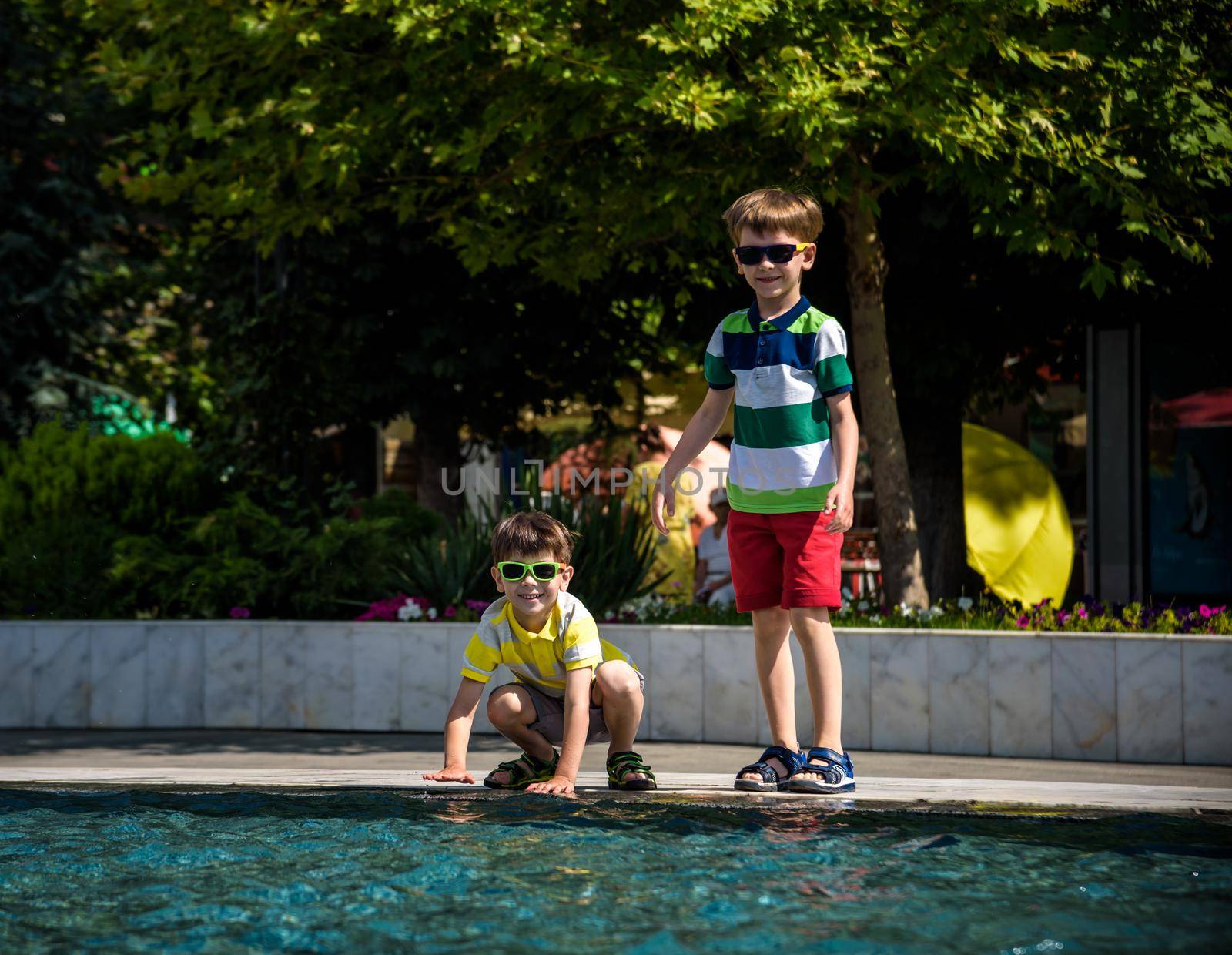 Group of happy children playing outdoors near pool or fountain. Kids having fun in park during summer vacation. Dressed in colorful t-shirts and shorts with sunglasses. Summer holiday concept by Kobysh