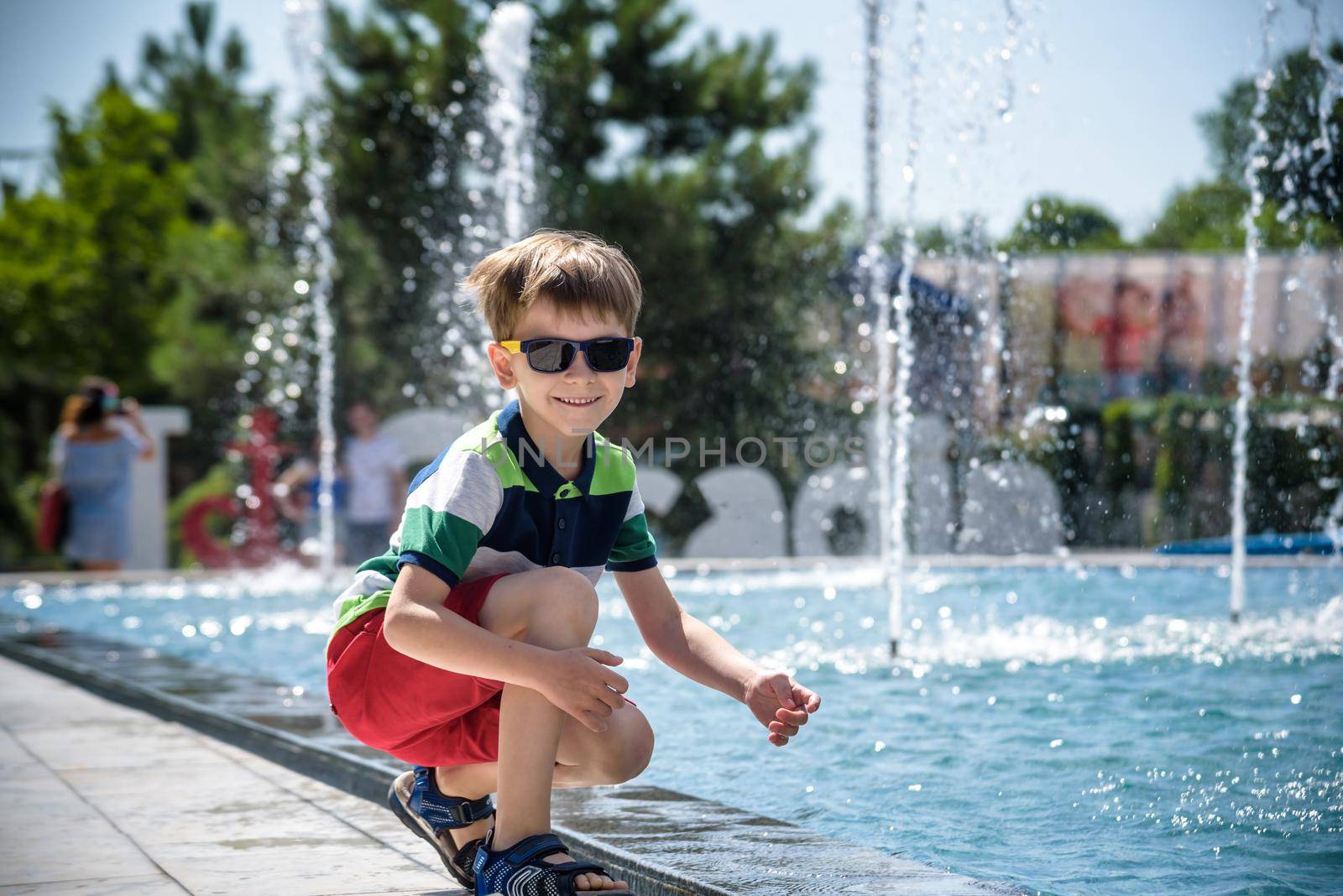 Little boy plays in the square near pool with water jets in the fountain at sunny summer day. Active summer leisure for kids in the city.