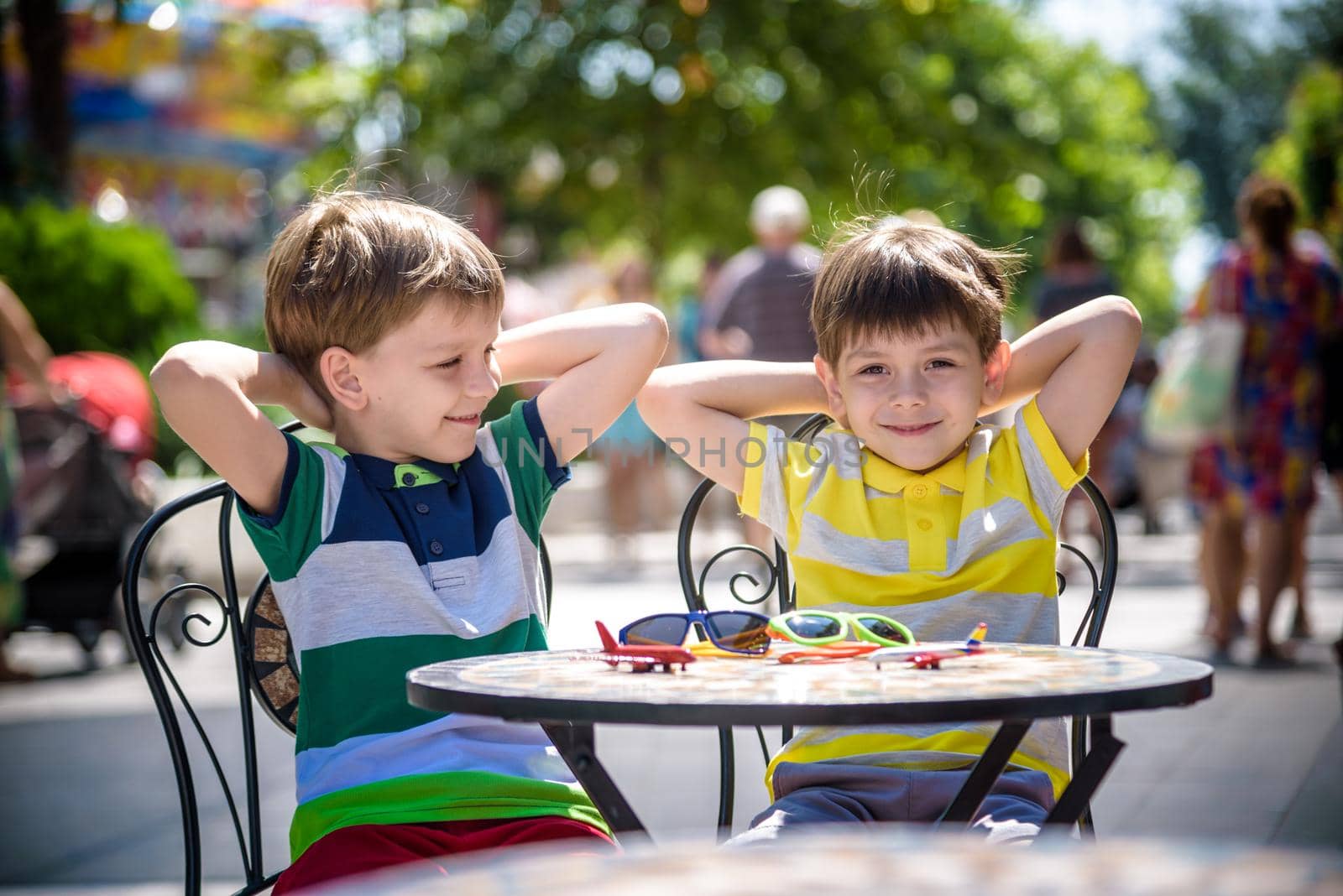 Two little kid boys waiting on table for healthy breakfast in hotel restaurant or city cafe. Child sit on comfortable chair with hands up, relaxed, enjoy his vacation. Summer holiday with children concept.