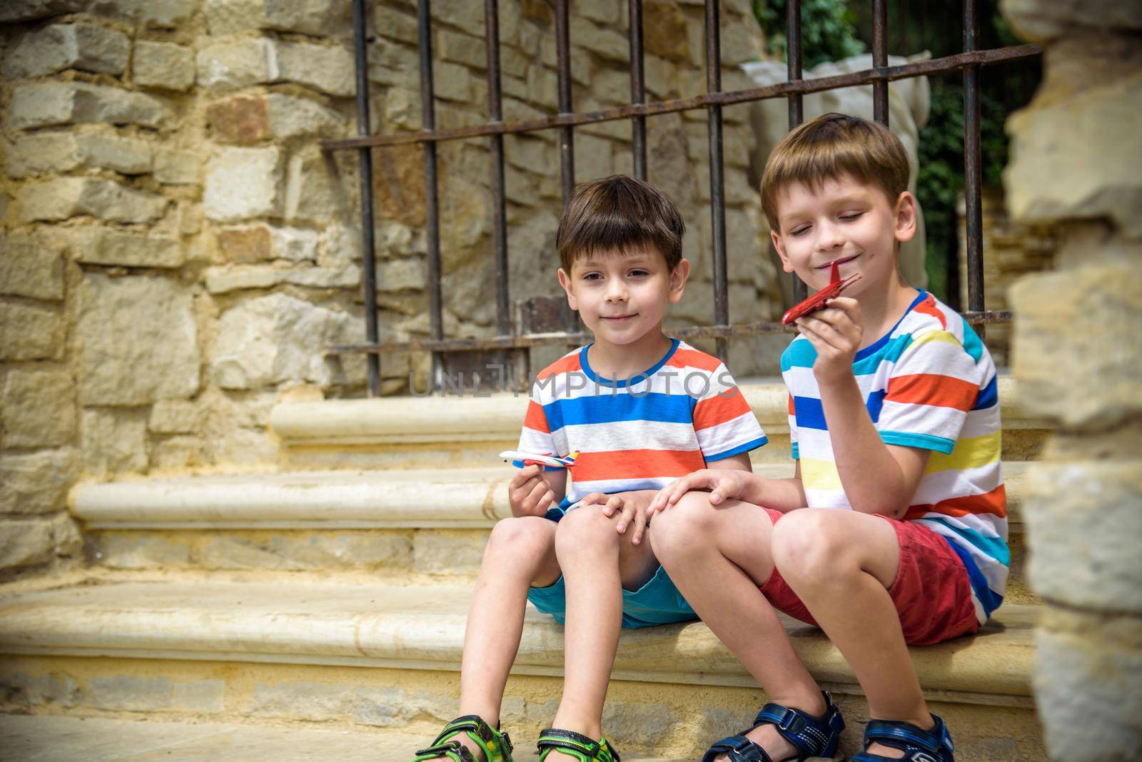 The children playing on the ruins of ancient building with metal gate an archaeological site of an ancient city. Two boys sitting and play with toy aircraft plane. Travel concept.