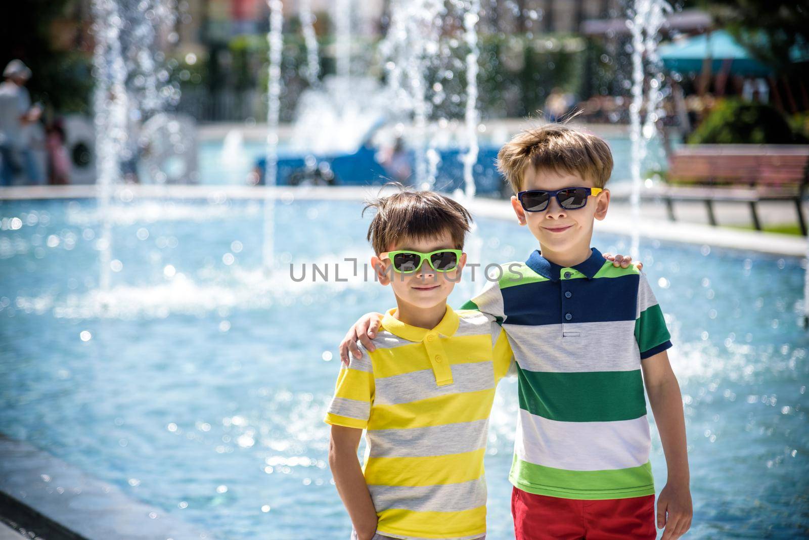 Group of happy children playing outdoors near pool or fountain. Kids embrace show thumb up in park during summer vacation. Dressed in colorful t-shirts and shorts with sunglasses. Summer holiday concept by Kobysh