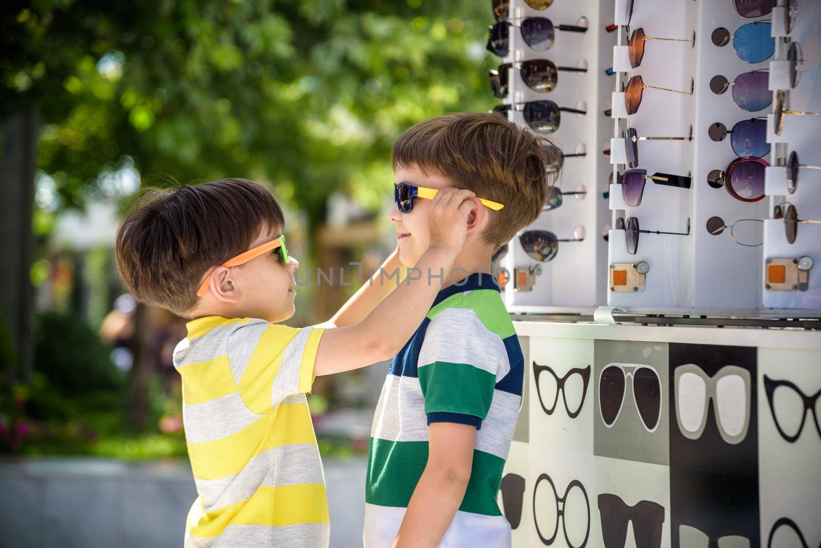 A child and a choice of sunglasses. Two little boys are standing in sun-proof glasses against the background of a shop window with glasses. Kid help choose each other. Sunglasses sale during summer vocation by Kobysh