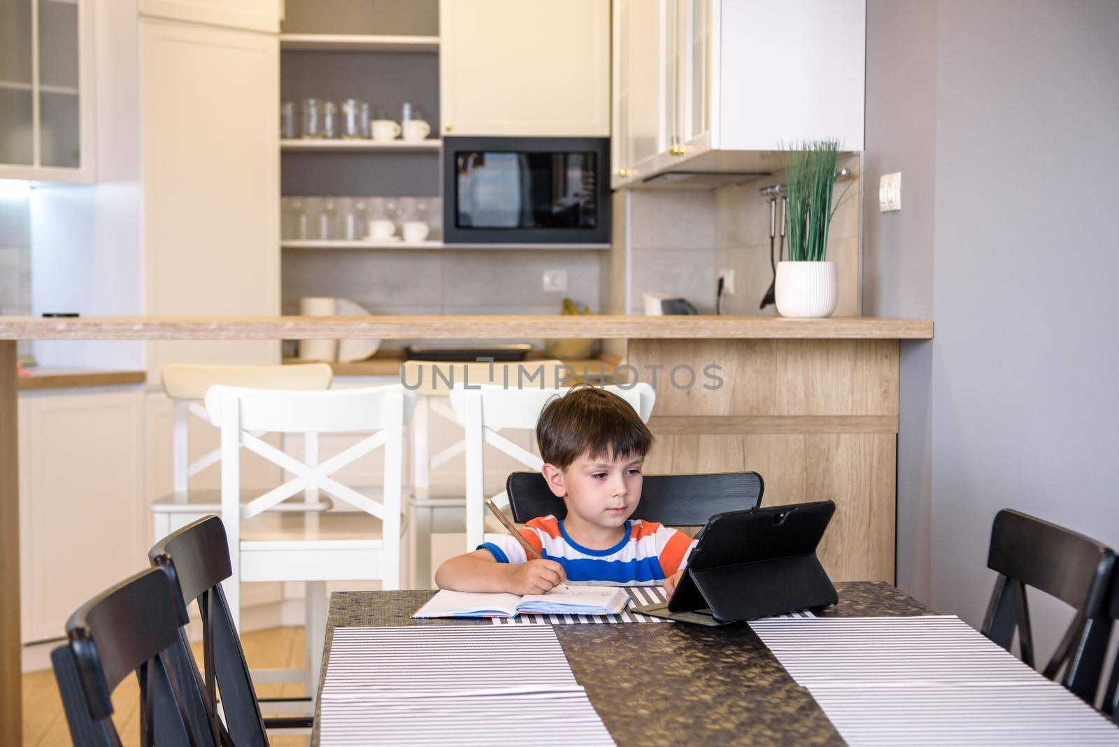 Smart preteen schoolboy doing his homework with digital tablet at home. Child using gadgets on his kitchen to study. Modern education and learning for kids.