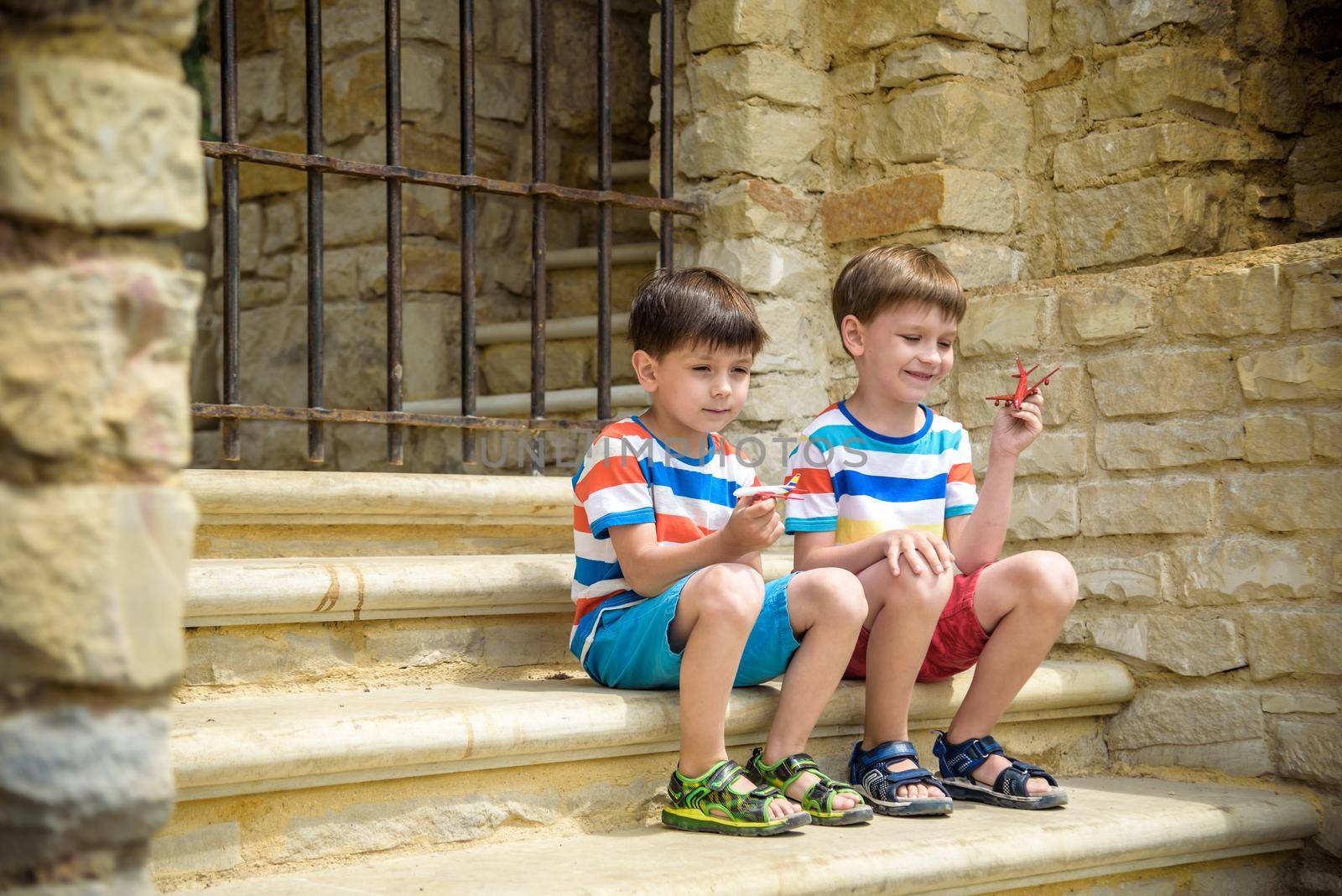 The children playing on the ruins of ancient building with metal gate an archaeological site of an ancient city. Two boys sitting and play with toy aircraft plane. Travel concept.