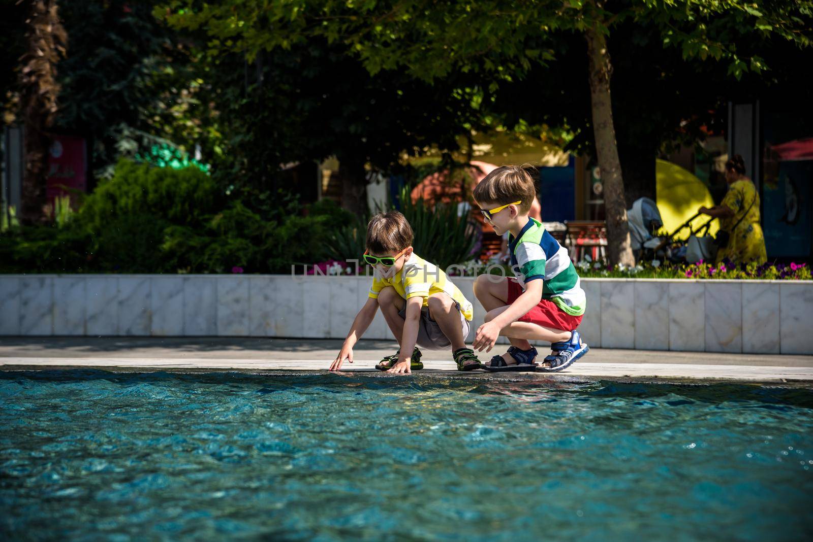 Group of happy children playing outdoors near pool or fountain. Kids having fun in park during summer vacation. Dressed in colorful t-shirts and shorts with sunglasses. Summer holiday concept.