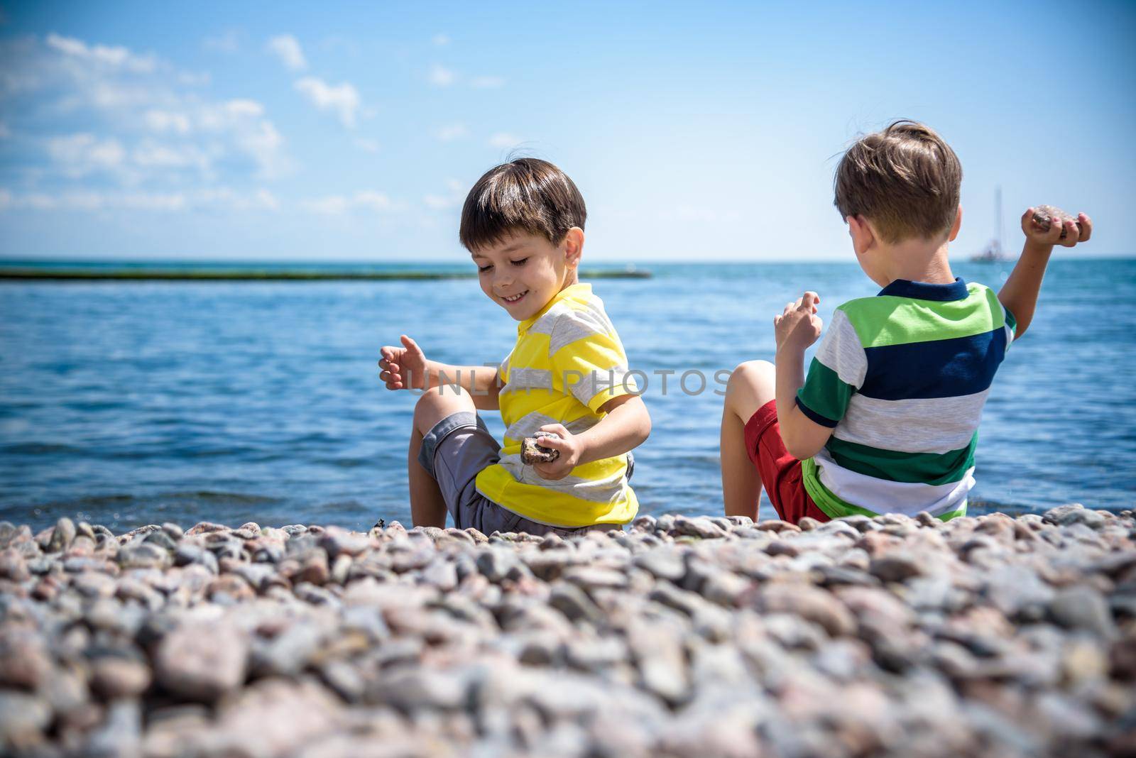 Two Caucasian baby boys is sitting at the beach. Kids in pants with his back sitting on the sea on a rocks pebbles. Sea vacations on with kids.