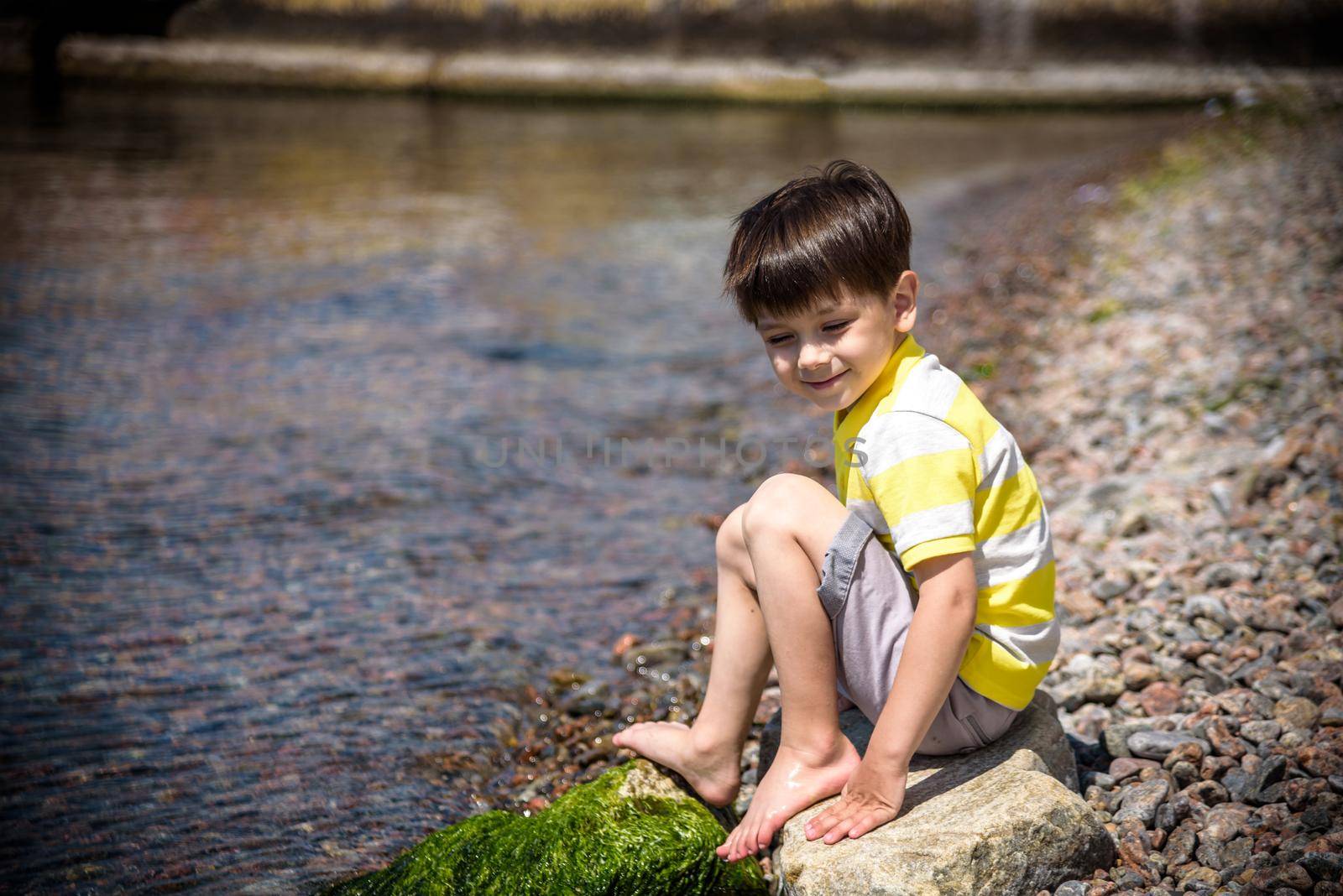 Caucasian baby boy is sitting at the beach. Cute child is playing with roks and shellfishes muscle-fish on the sand in summer day. Copyspace, close up, outdoor.