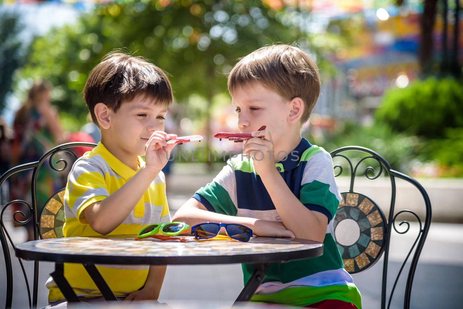 Two little kid boys waiting on table for healthy breakfast in hotel restaurant or city cafe. Child sit on comfortable chair play with toy aircraft, relaxed, enjoy their vacation. Summer holiday with children concept.