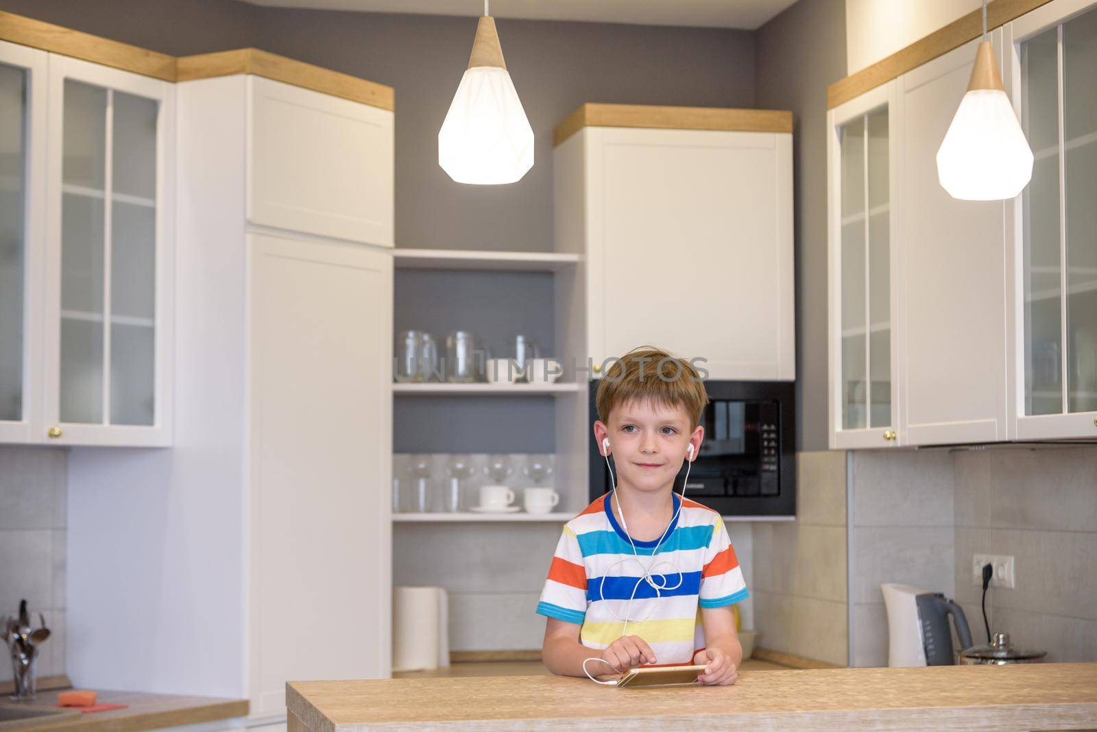 Little boy sitting at kitchen table holding a phone in his hands with earphones while looking to screen, messaging online using Wi-Fi having astonished expression.