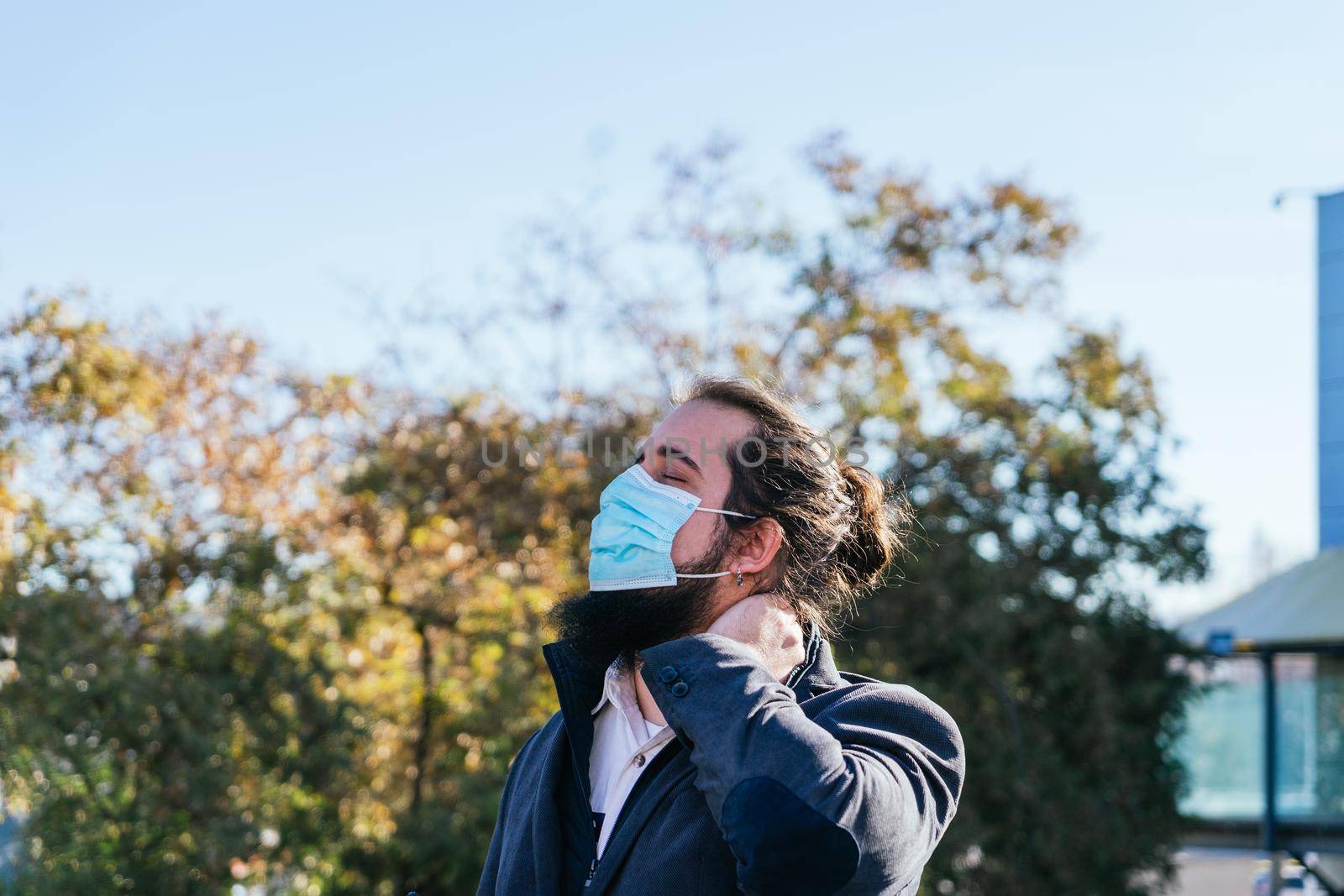 Young businessman with neck pain from work. He massages his neck near his offices on the street. Young entrepreneur with long hair and beard, brown, on a sunny day.