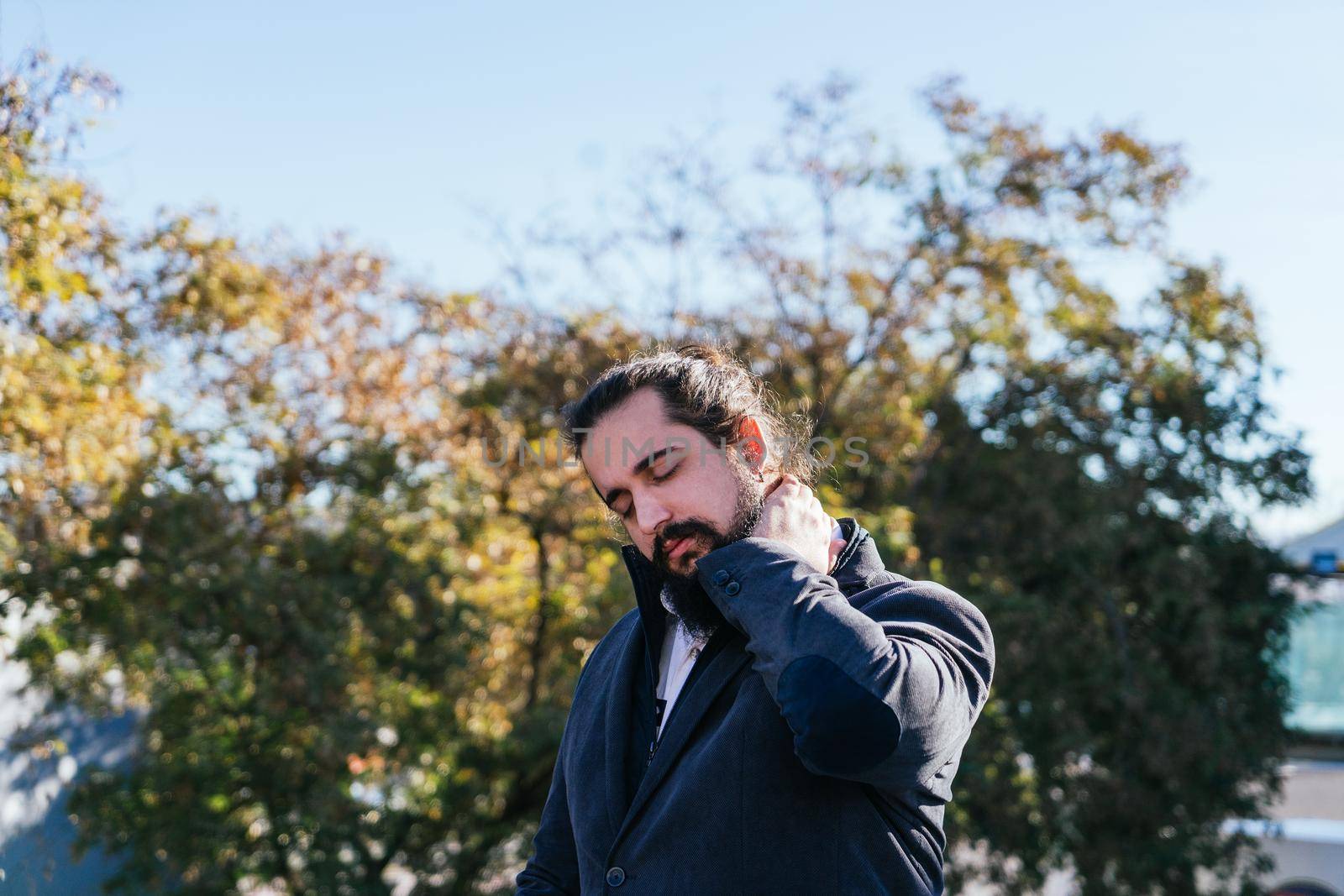 Young businessman with neck pain from work. He massages his neck near his offices on the street. Young entrepreneur with long hair and beard, brown, on a sunny day.