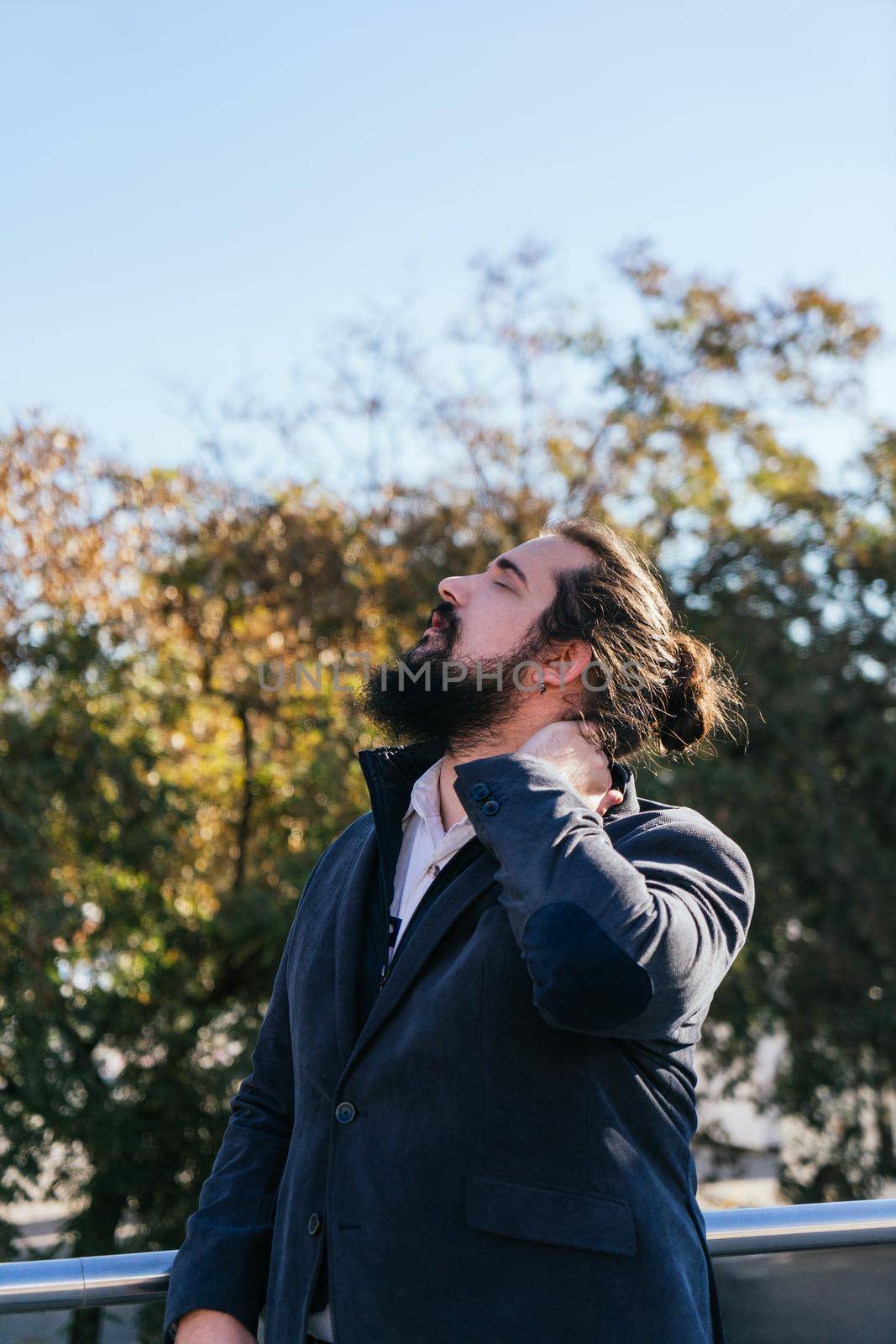 Young businessman with neck pain from work. He massages his neck near his offices on the street. Young entrepreneur with long hair and beard, brown, on a sunny day.