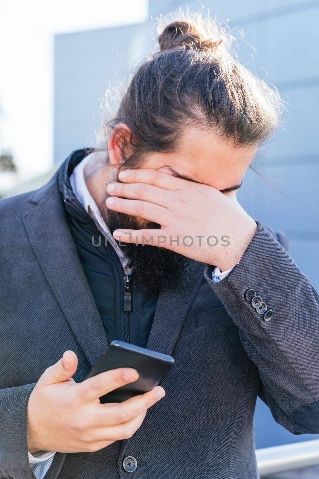 young businessman with long hair and beard, brown, laments because he receives bad news from his smartphone. He is outside near his offices during his work break.