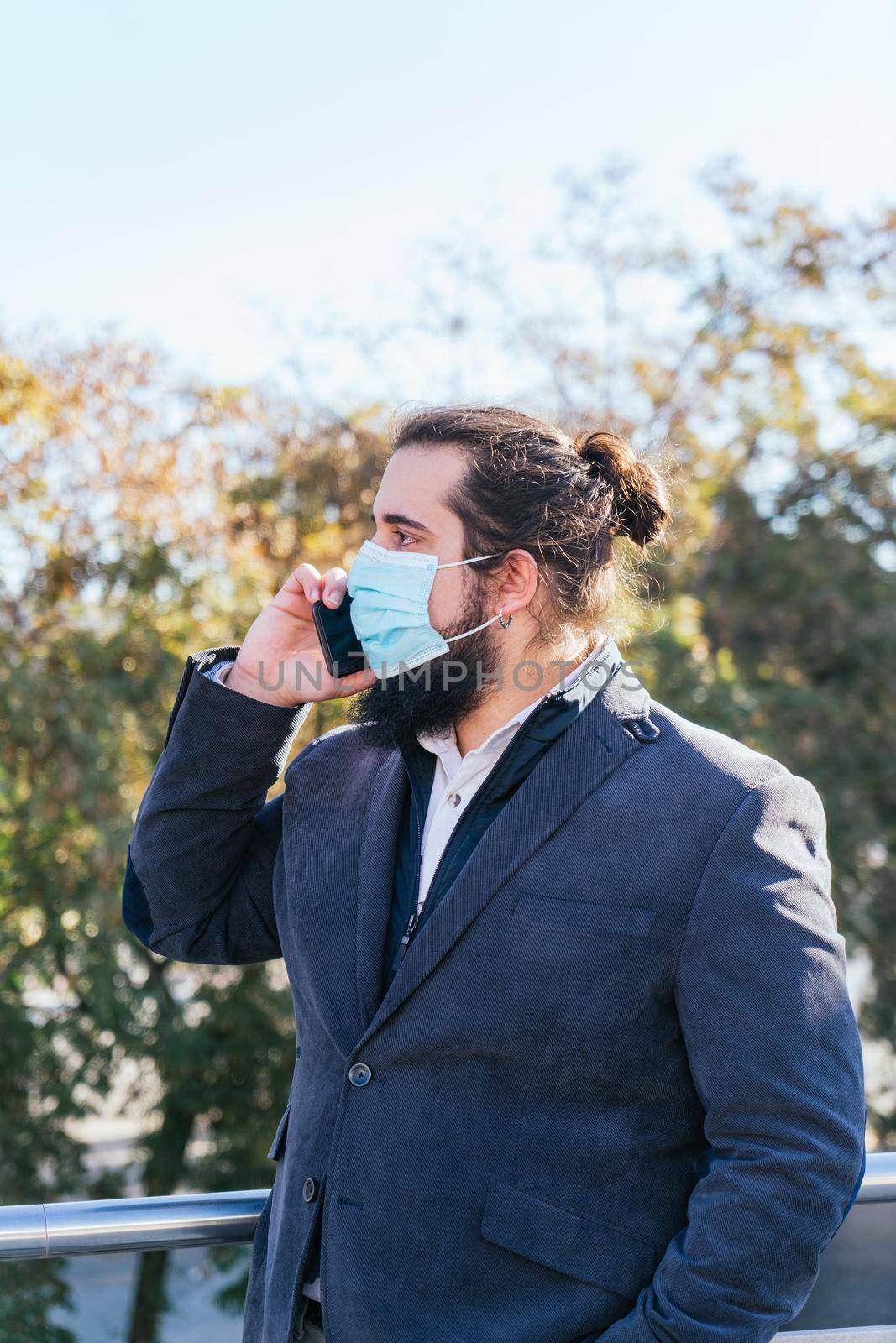 Young businessman with long hair and beard calling during his work break, on the street next to the offices. Vertical photo on a sunny and clear day. SIze view