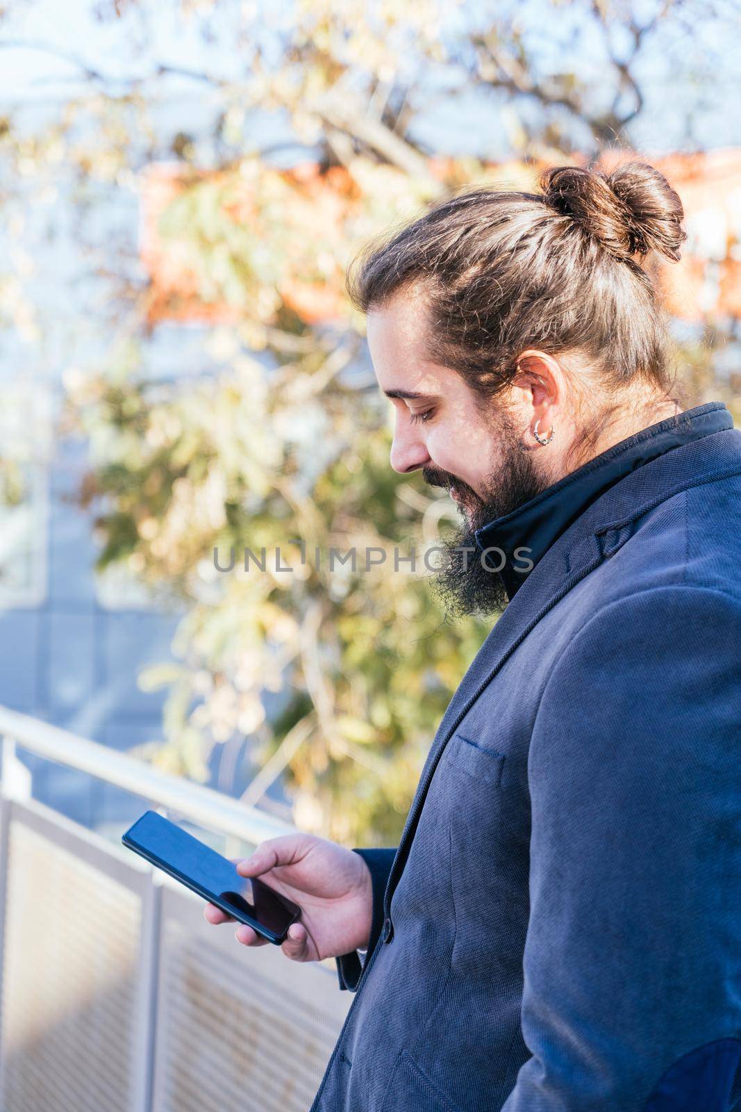 Young businessman with long hair and beard looking smartphone during his work break, on the street next to the offices. Vertical photo on a sunny and clear day. Size view.