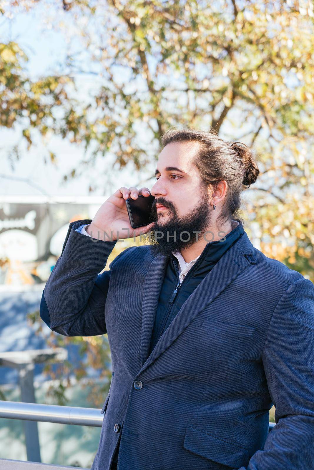 Young businessman with long hair and beard calling during his work break, on the street next to the offices. Vertical photo on a sunny and clear day.